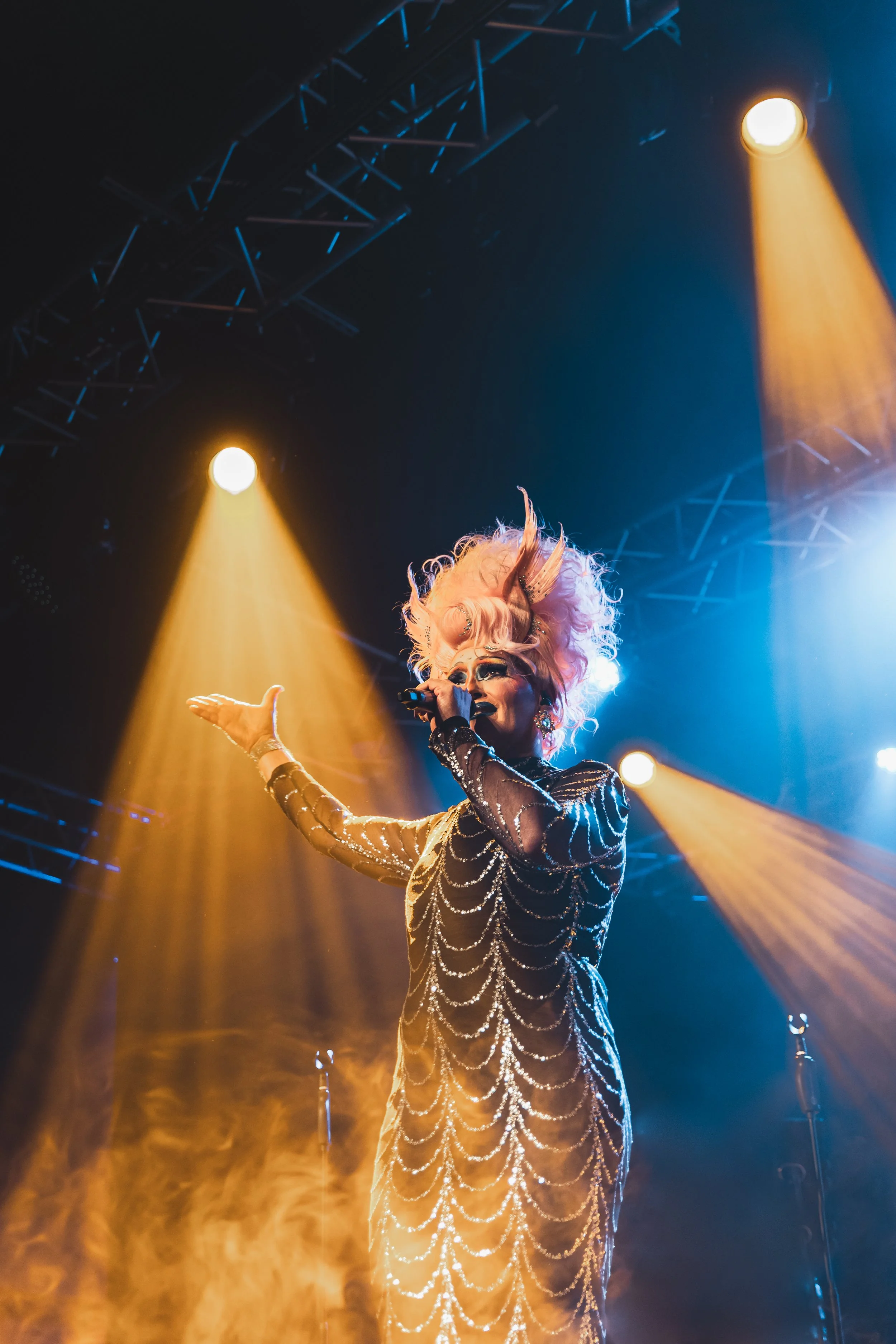 Performer with elaborate pink hair and glamorous makeup singing into a microphone on stage with bright orange and blue spotlights.