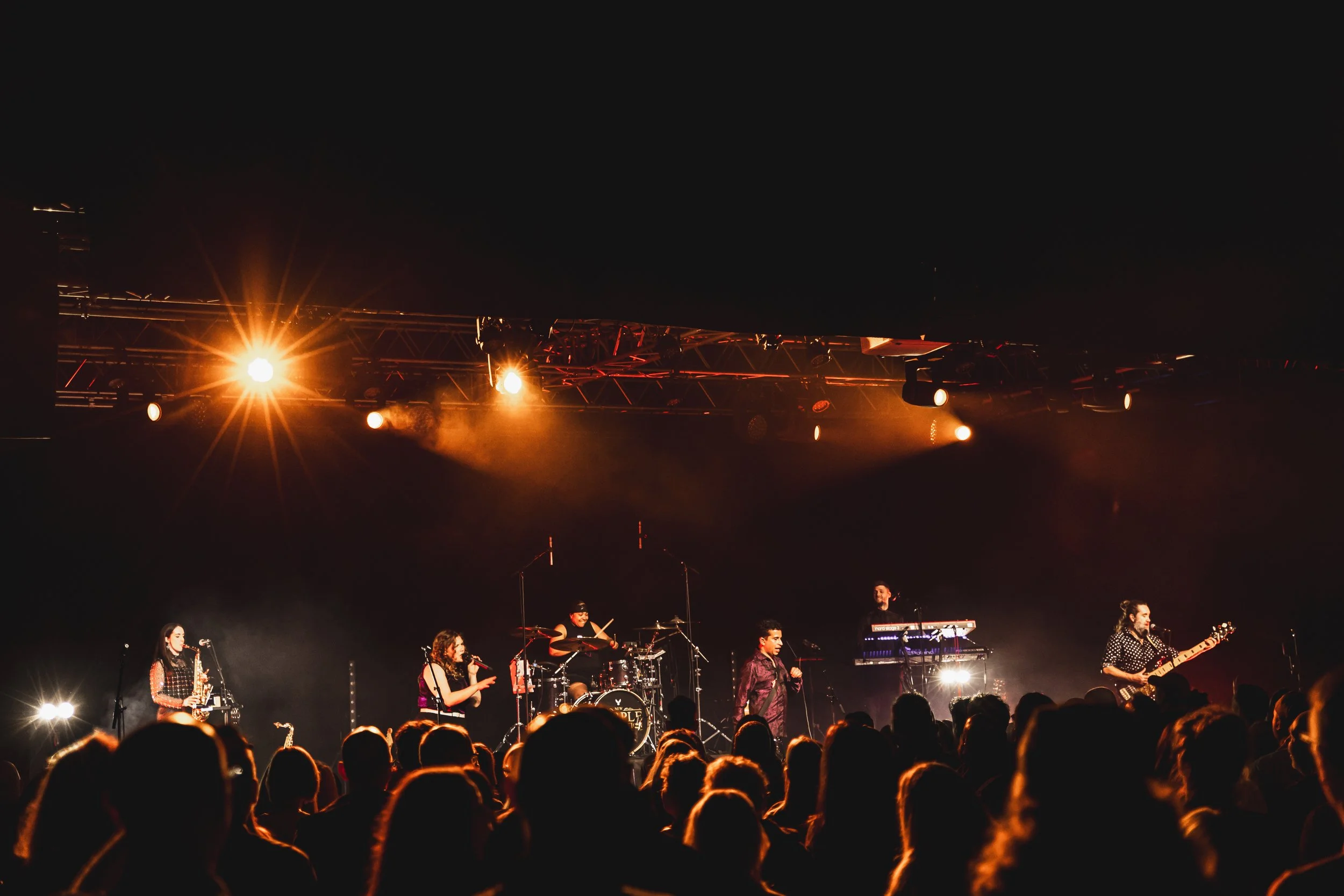 Prince tribute act, New Purple Celebration performing on stage at concert with bright stage lights and audience in foreground.