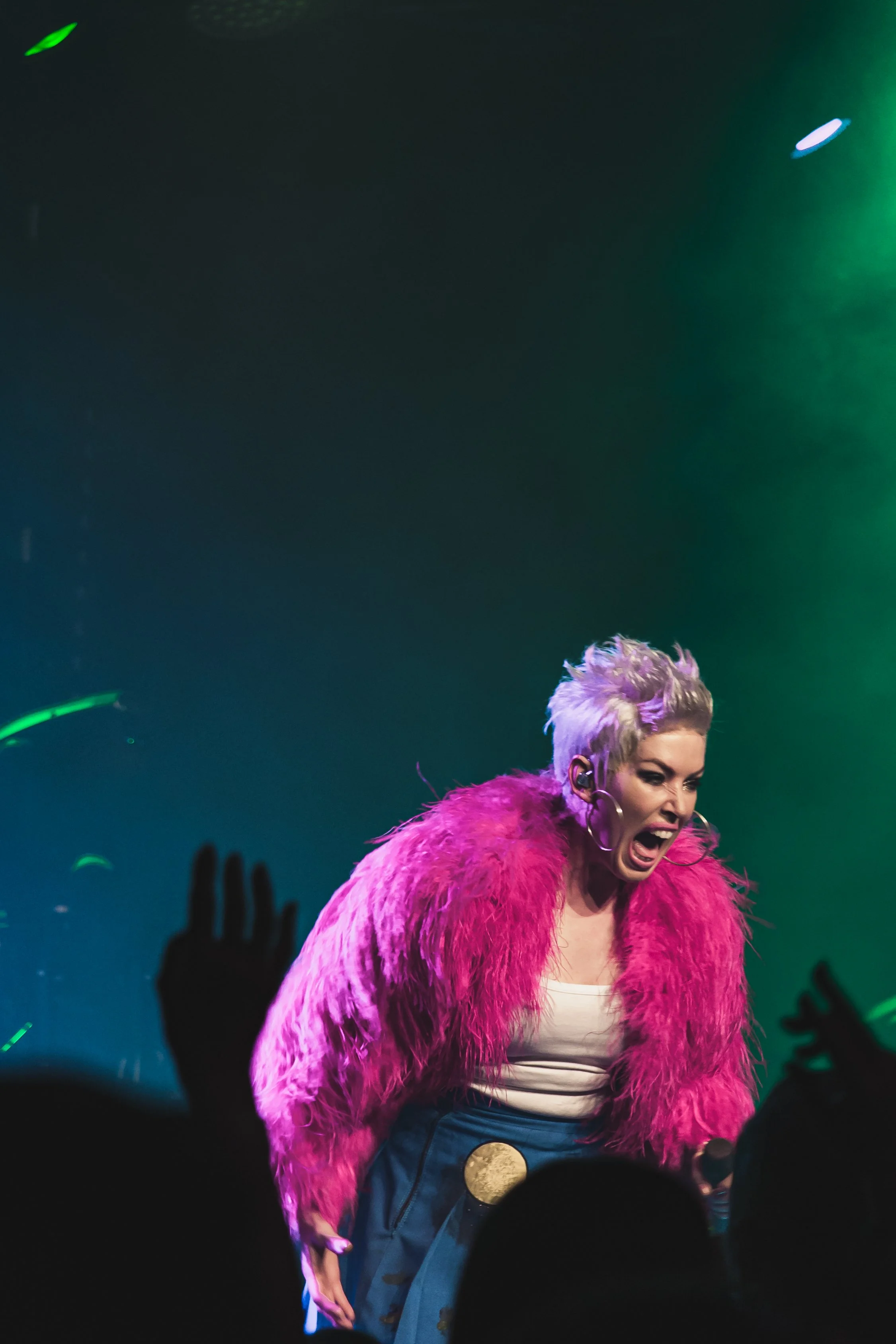 Performer with short, platinum blonde hair, wearing a bright pink fur coat, singing passionately on stage at a concert, with audience hands in the foreground and colorful stage lighting.