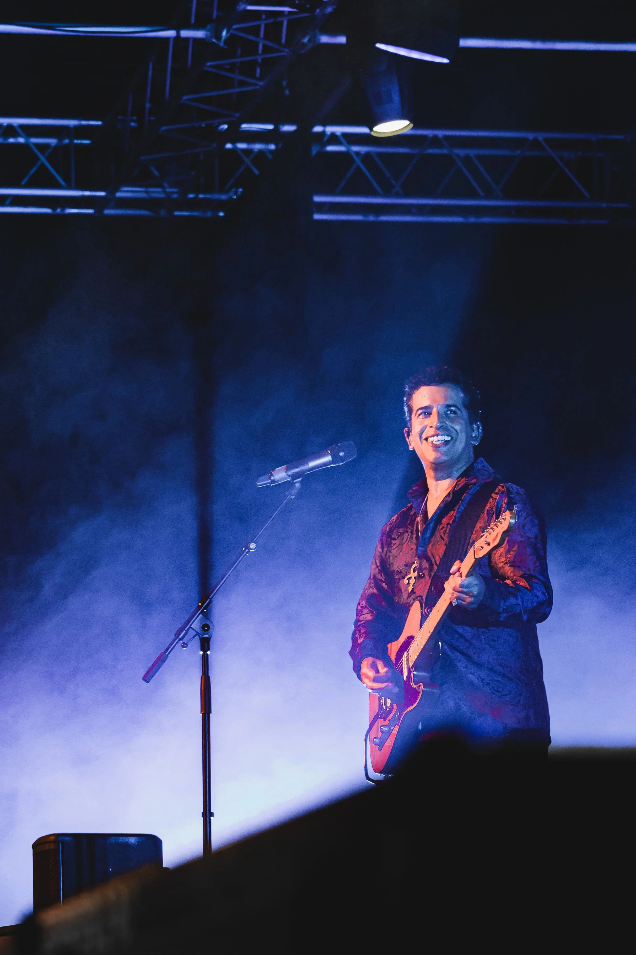 Prince tribute act, New Purple Celebration performing on stage with a guitar, smiling, illuminated by blue and white stage lights, with a microphone stand in front of him and stage equipment visible in the background.