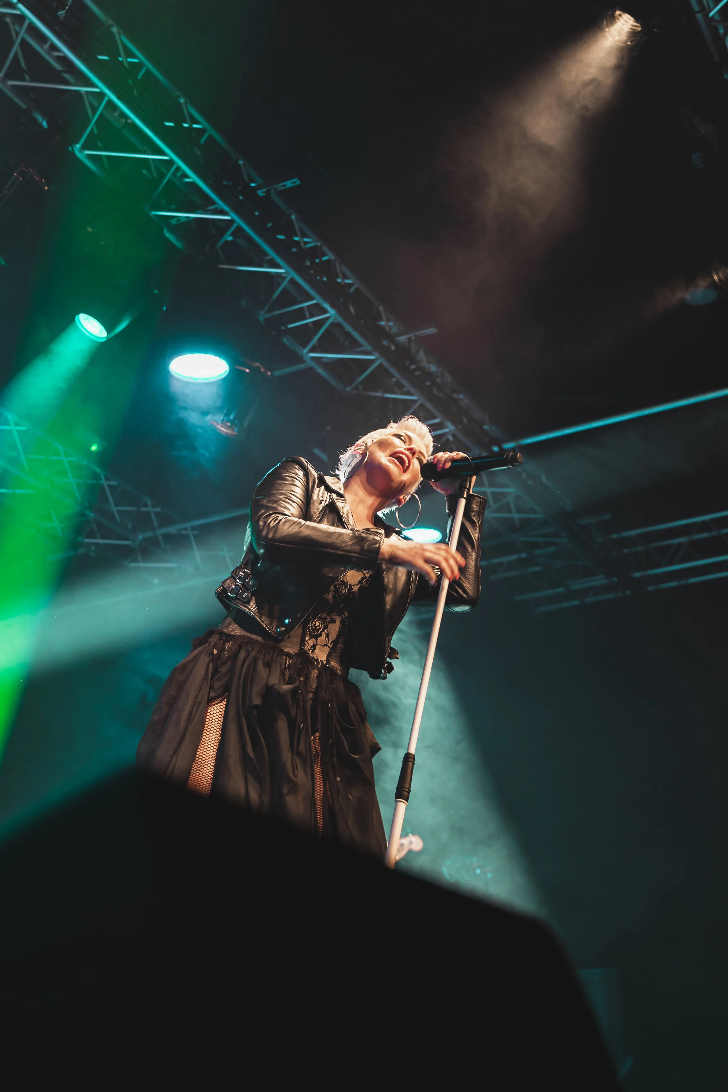 Female singer performing on stage with a microphone, stage lighting, and metal truss overhead.