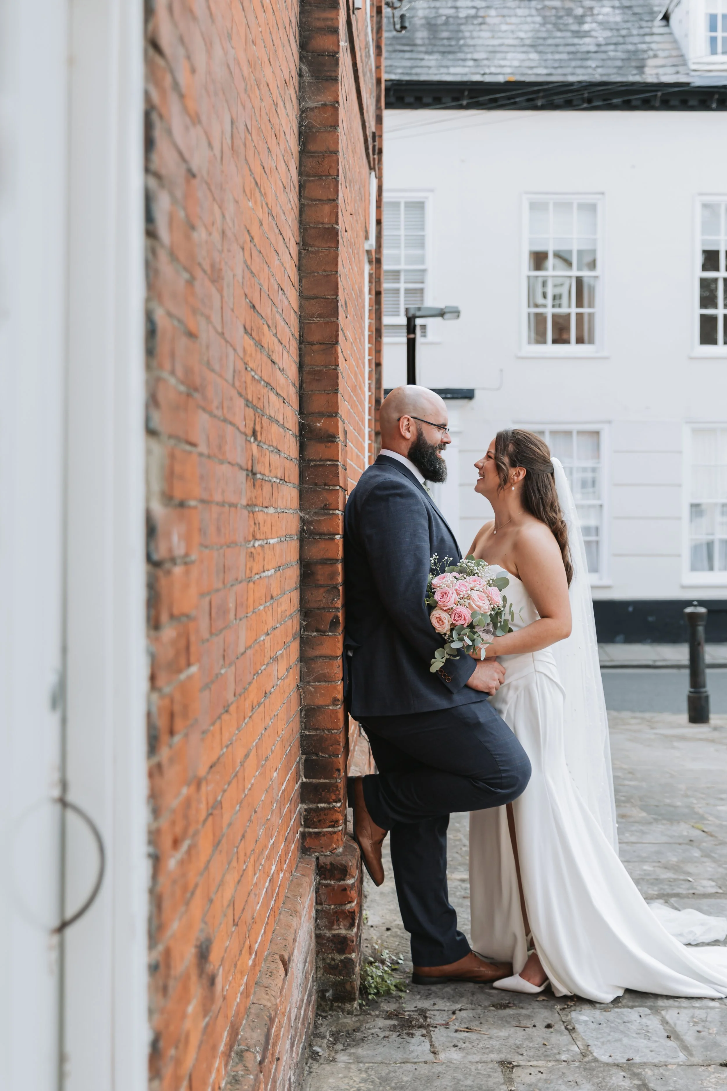 A bride and groom stand closely facing each other against a brick wall in an outdoor urban setting. The groom is dressed in a dark suit, and the bride is in a white wedding gown holding a bouquet of pink and white flowers. They are smiling and lookin