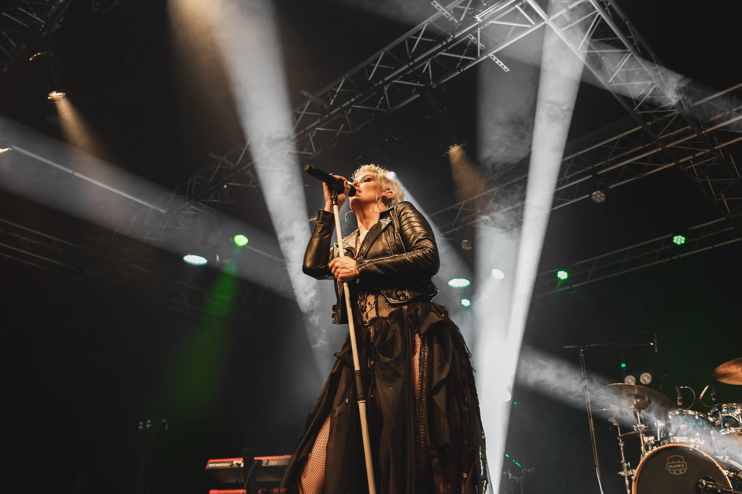 A female singer performing on stage with microphone, wearing a leather jacket and black skirt with lace details, illuminated by stage lights.