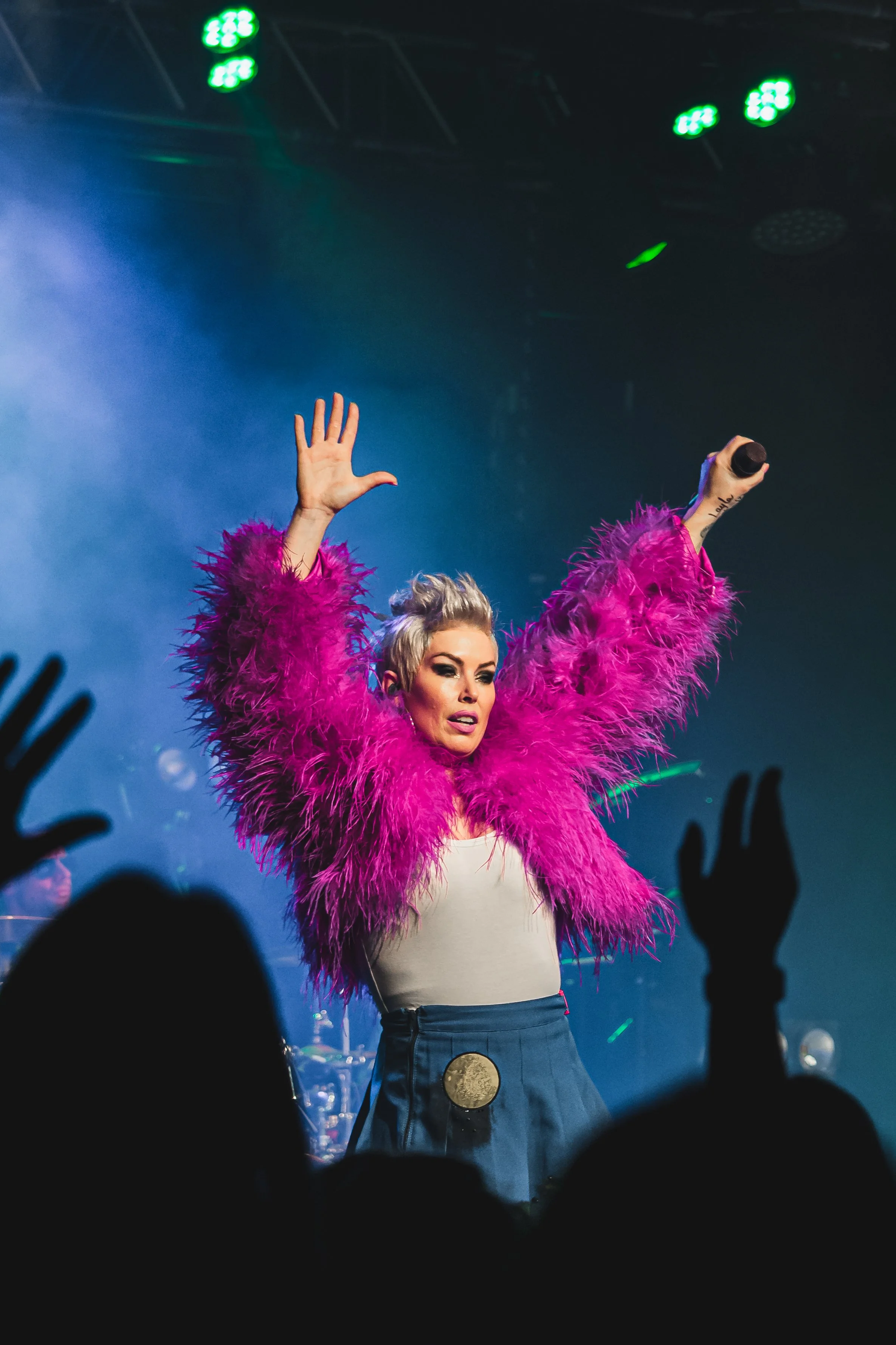 A female performer on stage wearing a bright pink feathered jacket and high-waisted blue pants, holding a microphone in one hand, with her other hand raised, in front of an audience.