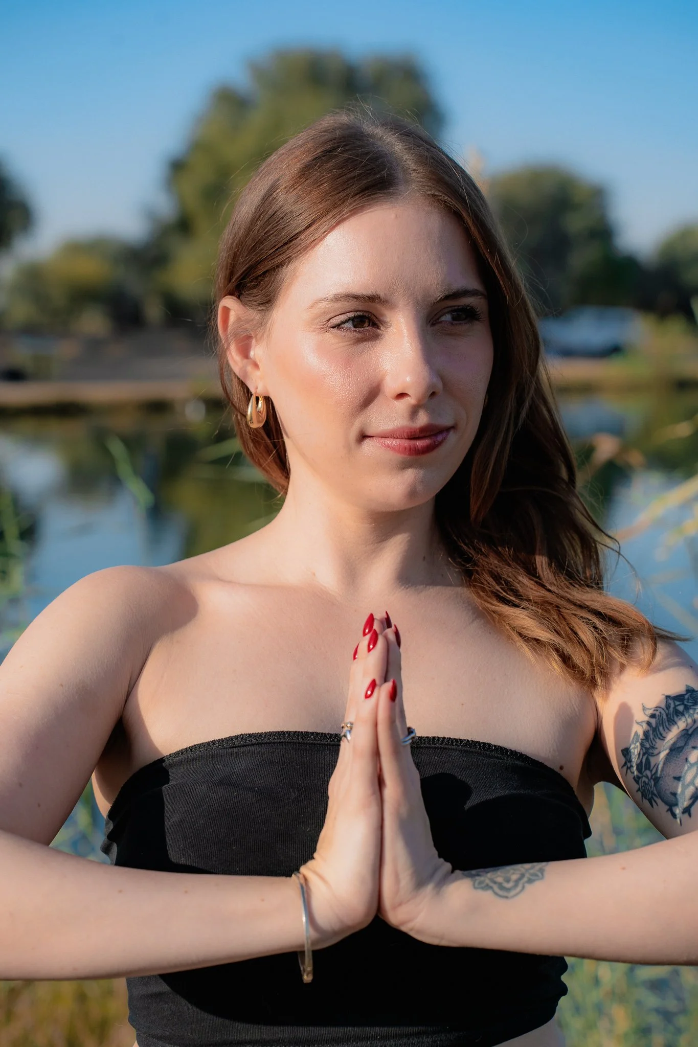 A young woman practicing yoga outdoors with her hands pressed together in a prayer position, standing near a body of water with trees in the background under a blue sky.