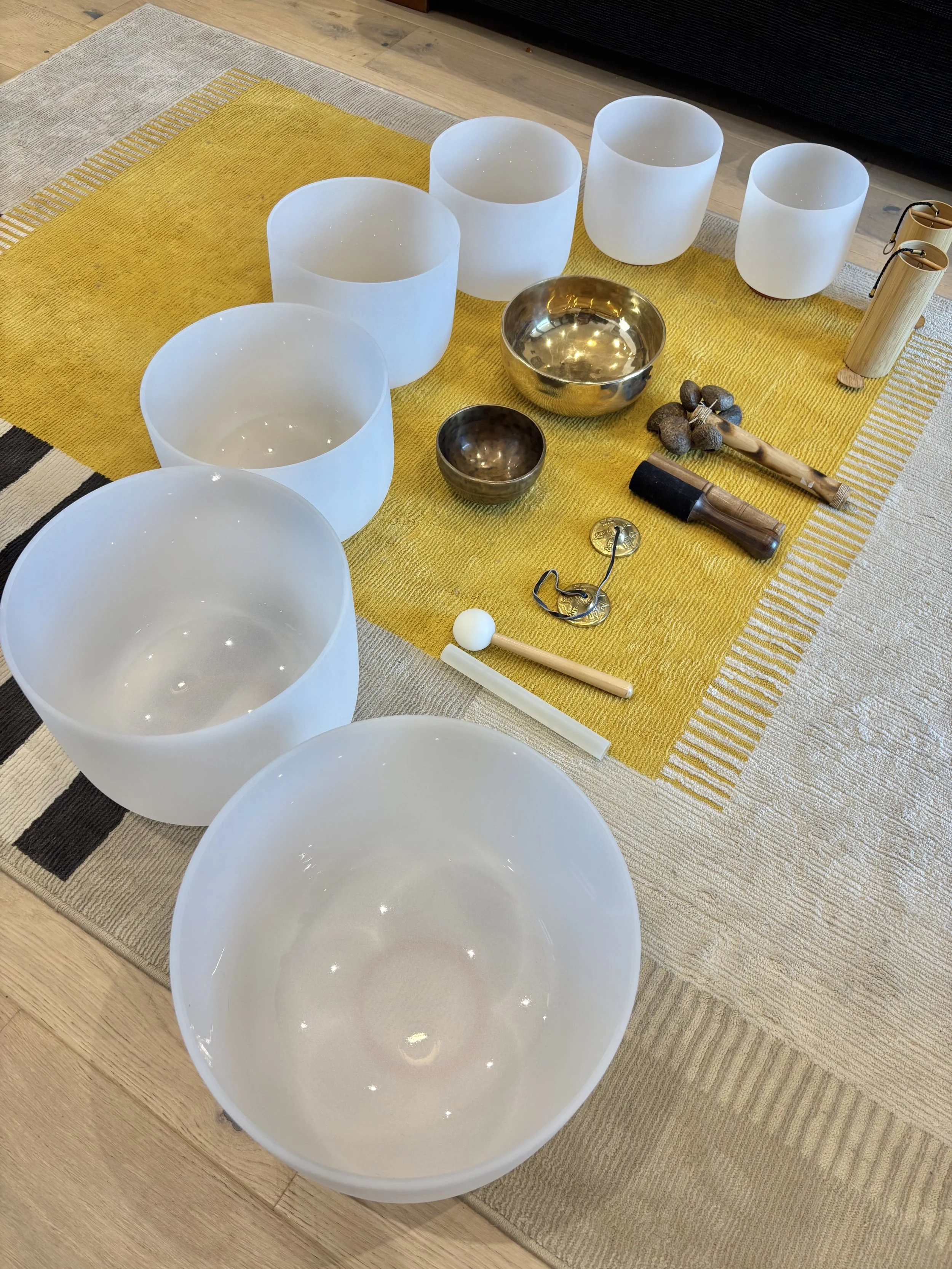 A set of six white crystal singing bowls arranged on a yellow and beige striped rug, with two smaller metallic bowls, a mallet, a small drum, and a tuning fork nearby.
