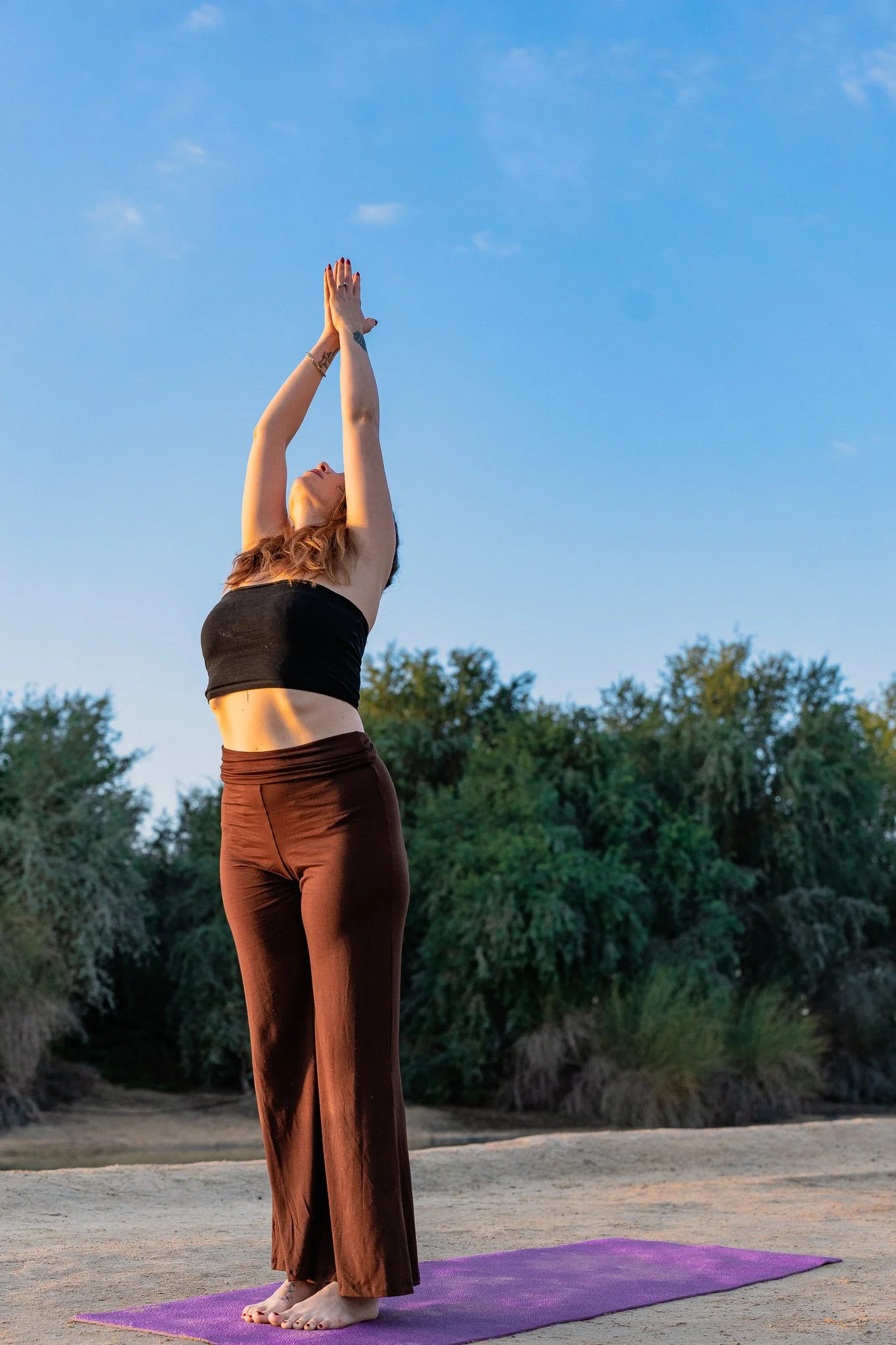 A woman practicing yoga outdoors on a purple mat, standing on a sandy surface with green trees in the background, raising her arms overhead with palms together, during late afternoon or early evening.