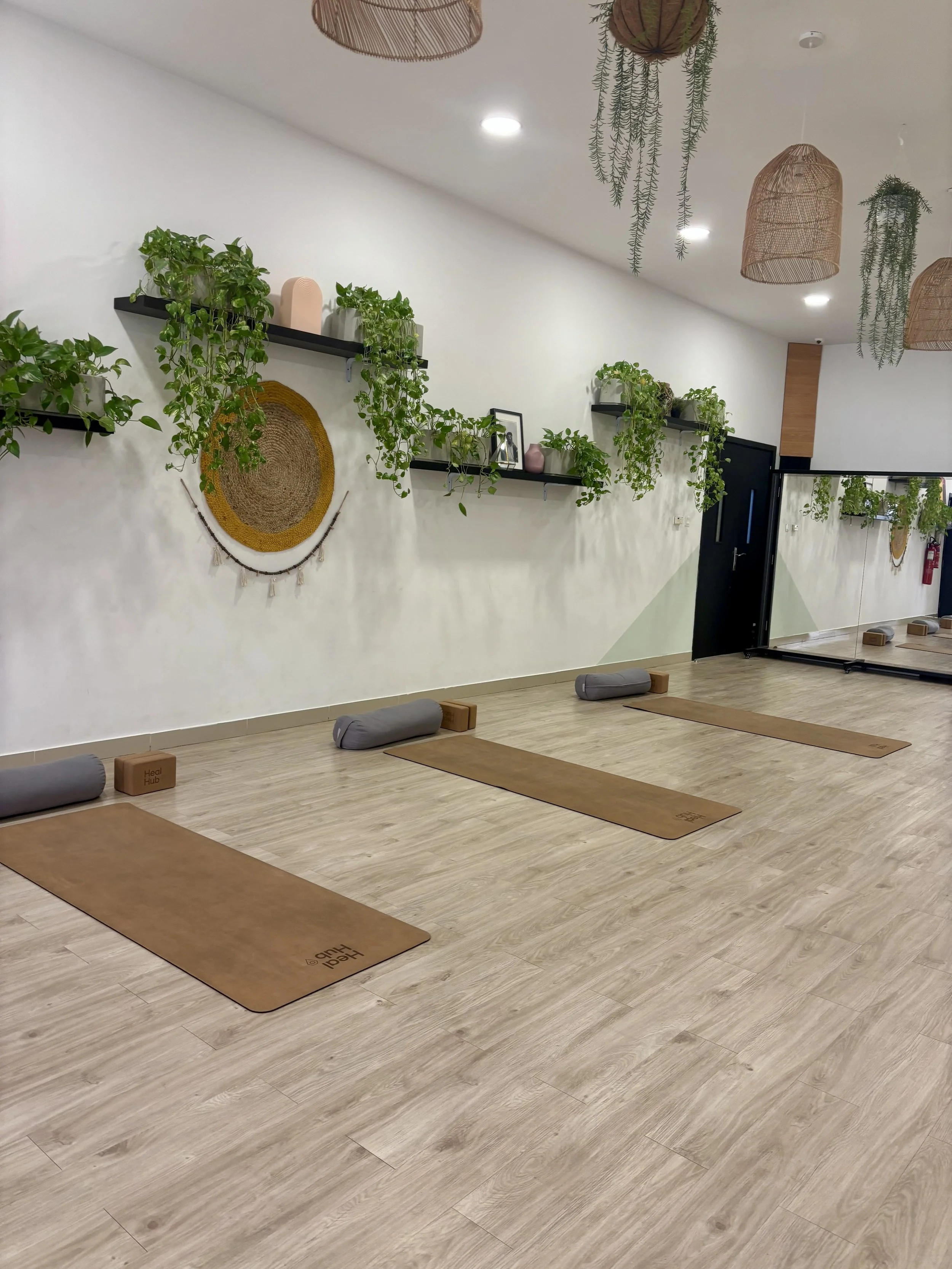 Yoga studio with wooden floor, yoga mats, bolsters, and blocks arranged in a spacious, well-lit room with greenery and decorative hanging baskets.