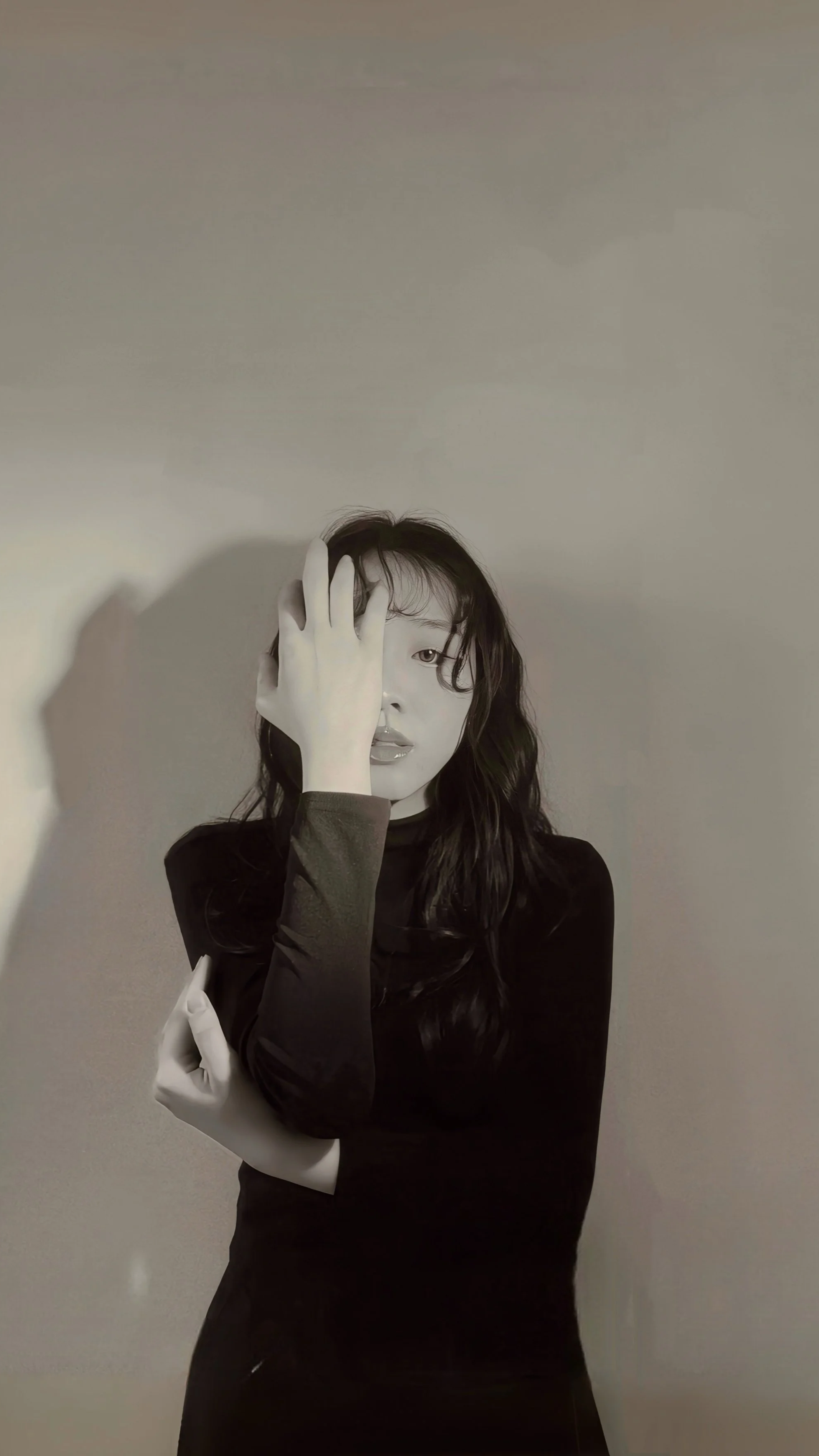 Black and white photo of a woman with long, wavy hair, partially covering her face with her hand, wearing a dark long-sleeve top, standing against a plain wall.