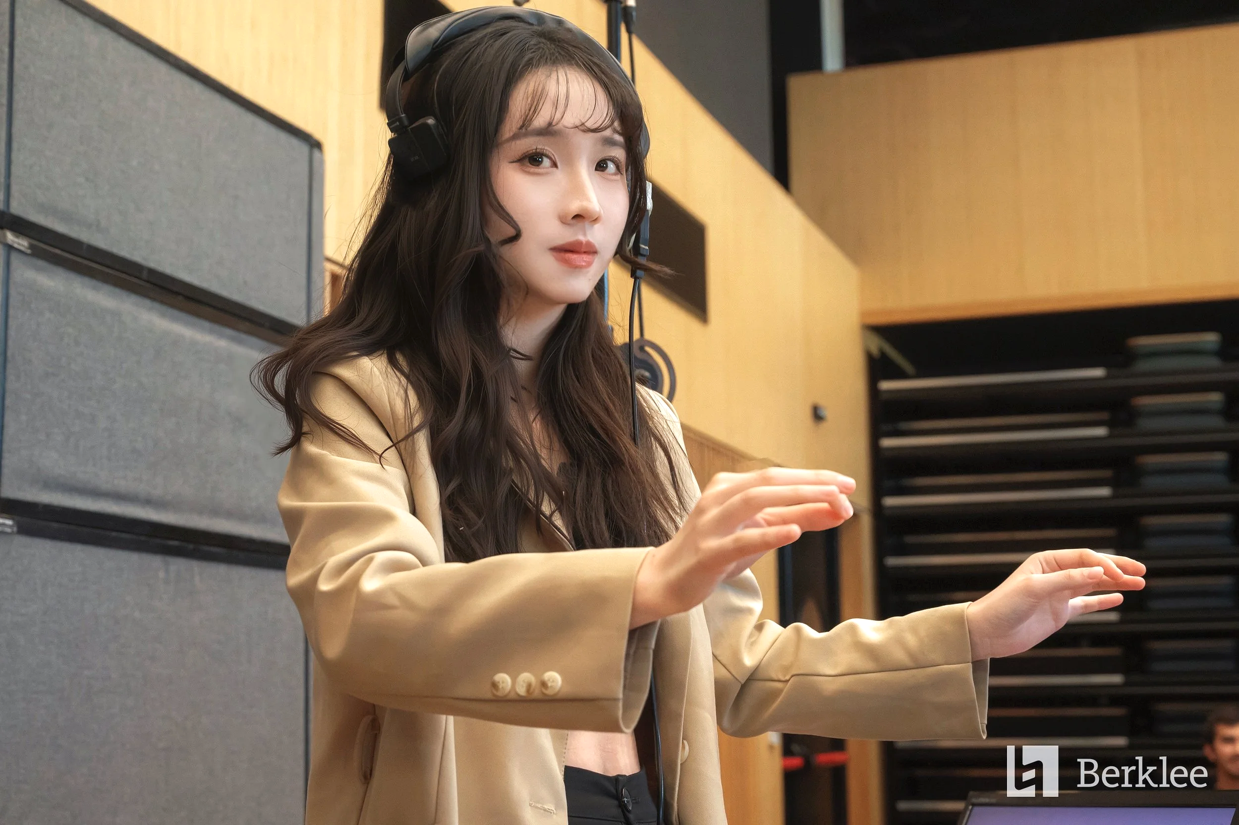 A young woman with long, wavy dark hair wearing a beige blazer, black top, and headphones in a recording studio, gesturing with her hands.