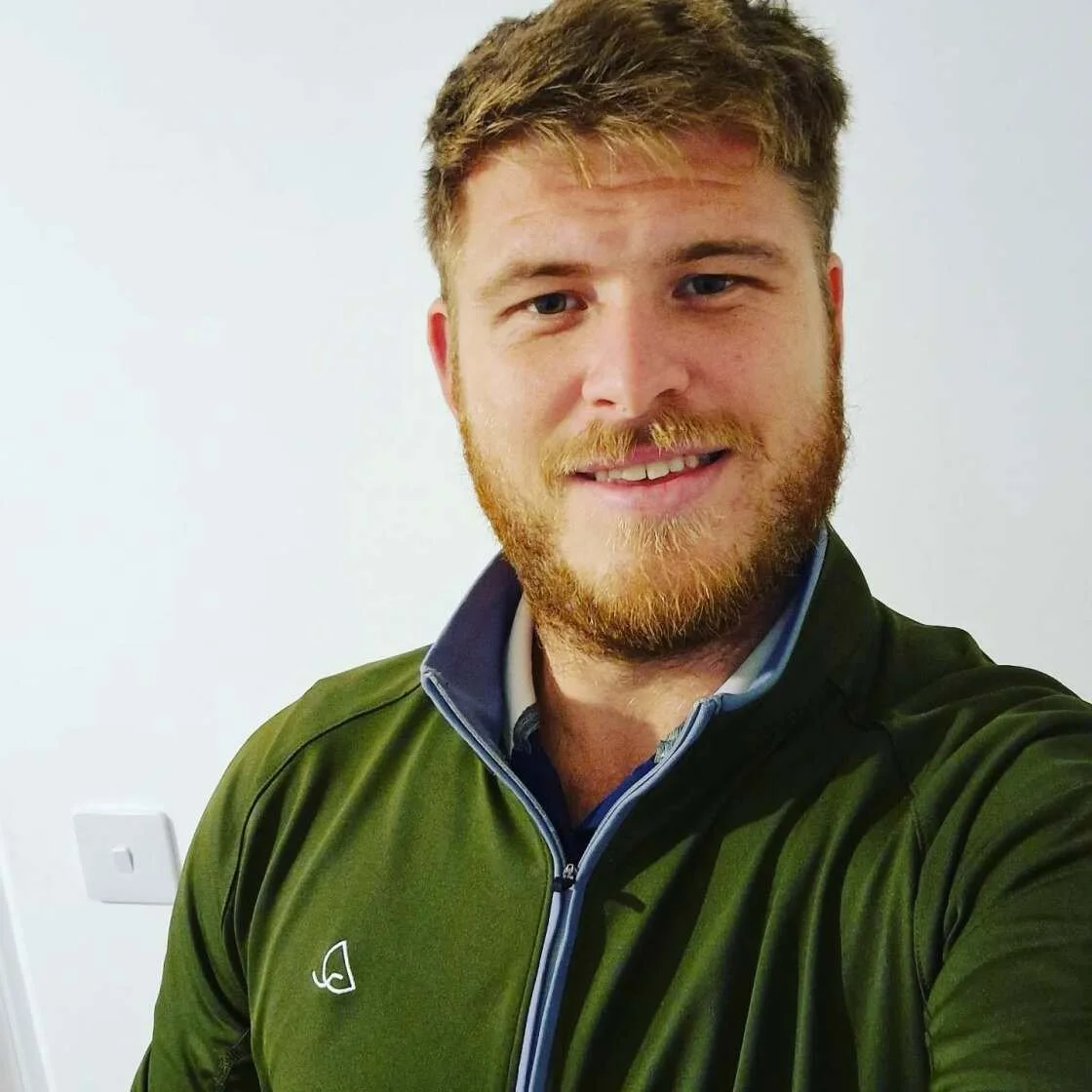 A young man with red hair and a beard smiling at the camera, wearing a green jacket with a zipper and a collar, standing against a plain light-colored wall.