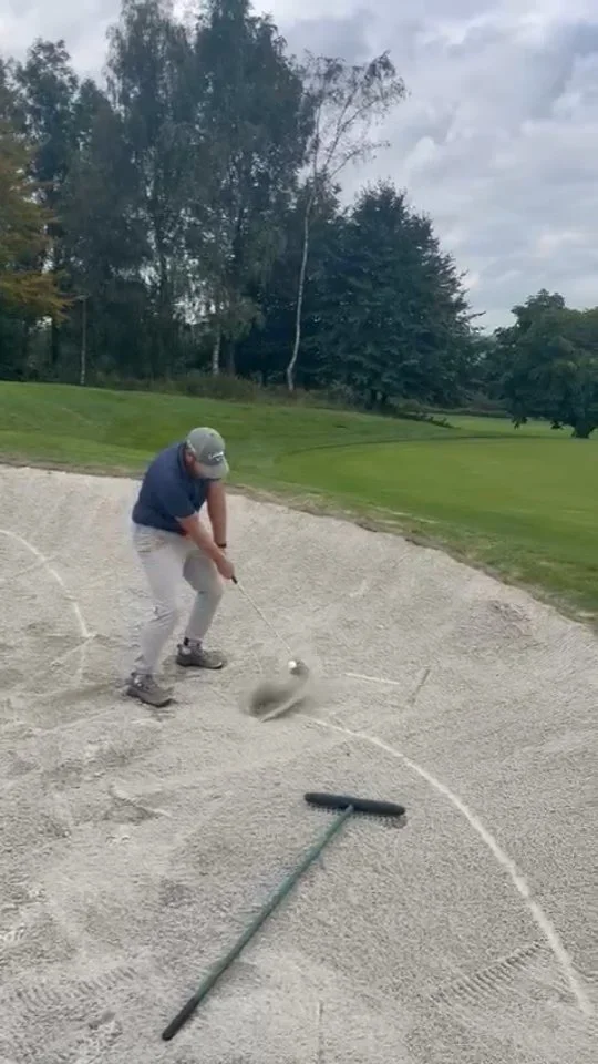 A man in a blue shirt, gray pants, and a gray cap hitting a golf ball out of a bunker on a golf course. A rake lies in the sand nearby, and trees and grass are visible in the background.