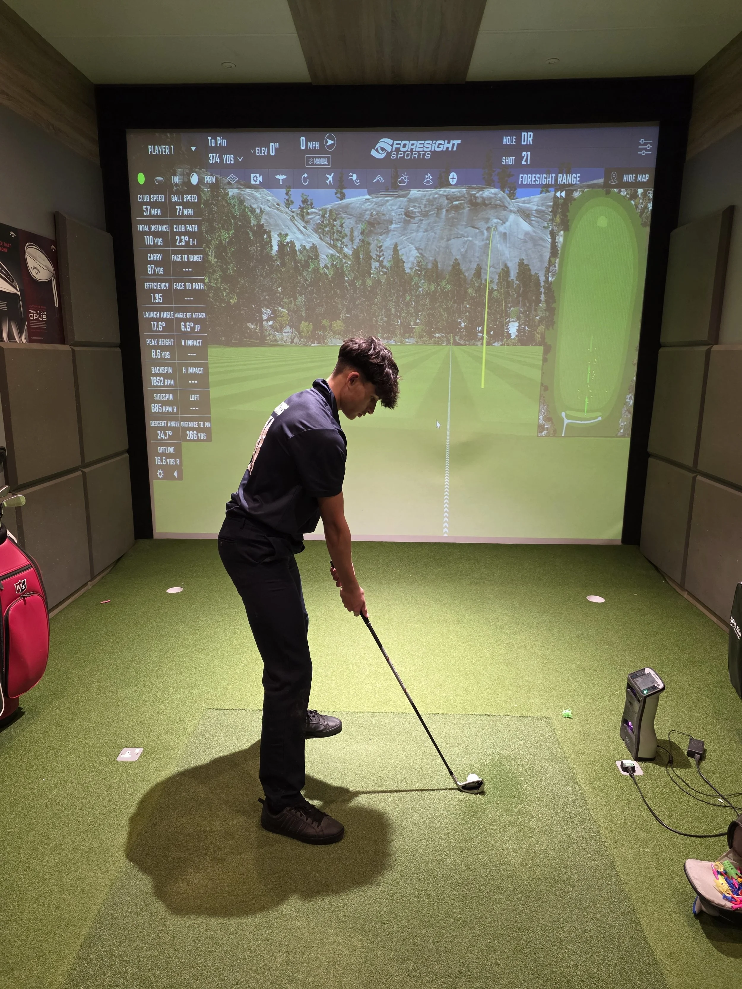 A young man in a black outfit is practicing golf swings inside a golf simulation room with a large screen showing a virtual golf course. The screen displays golf data and a scenic background of trees and mountains.