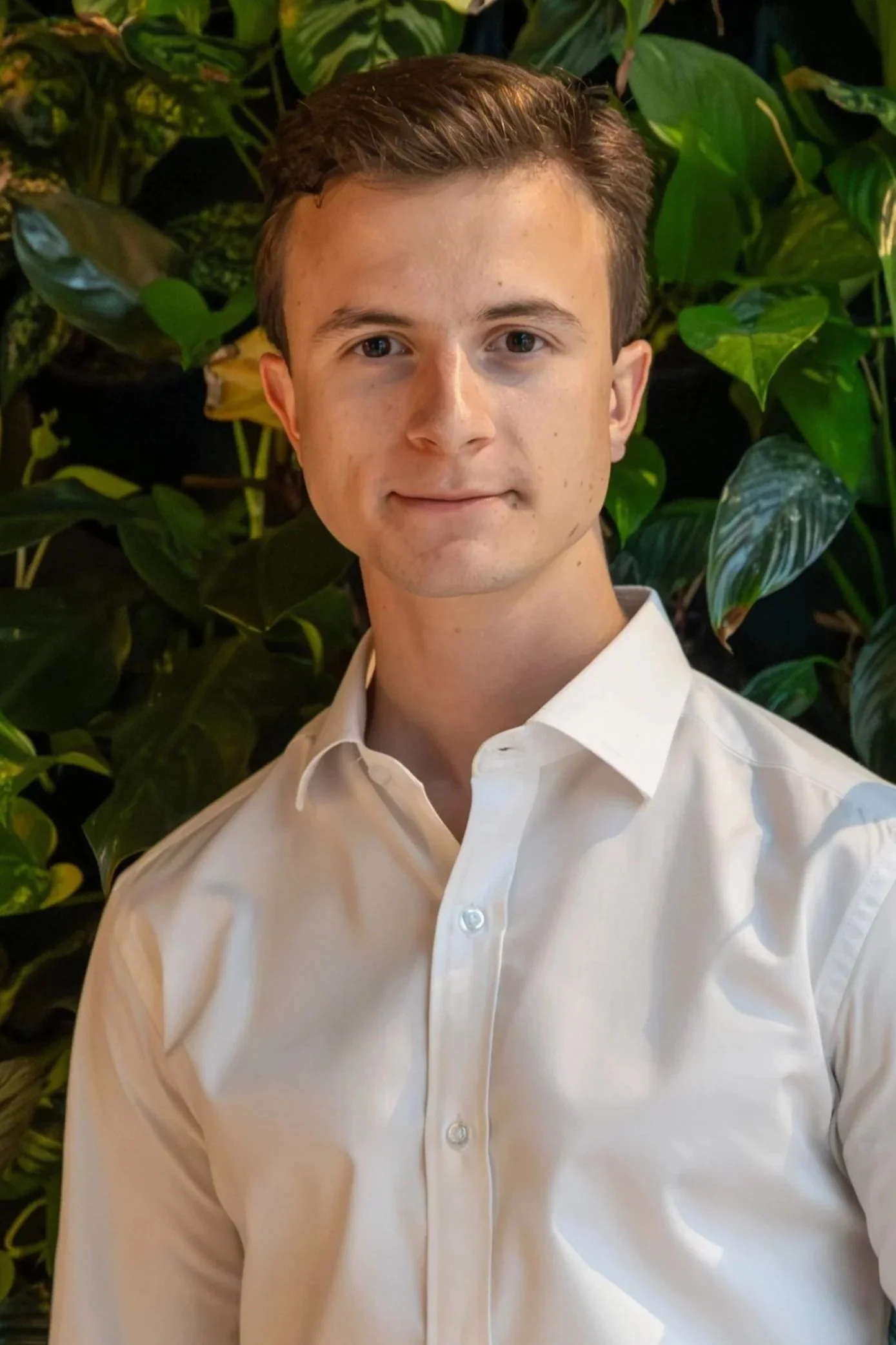 Portrait of a young man with short brown hair, wearing a blue dress shirt and dark blazer, standing in front of a plain gray background.