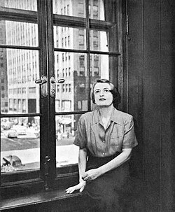 Ayn Rand leaning against a windowsill in an urban office, looking upwards with a thoughtful expression.