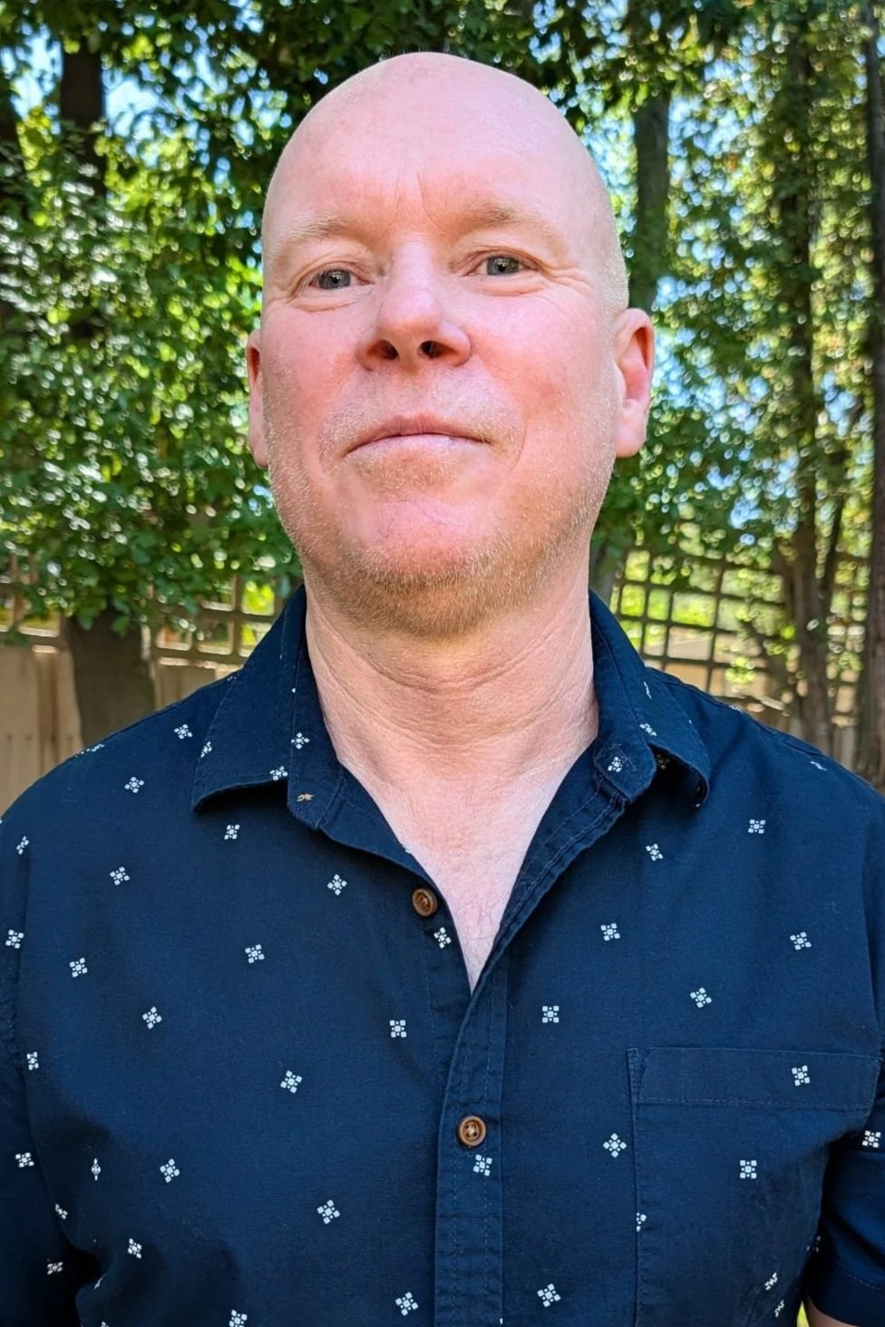 A man with a shaved head and light beard wearing a dark navy shirt with small white geometric patterns, standing outdoors with trees and a wooden fence in the background.