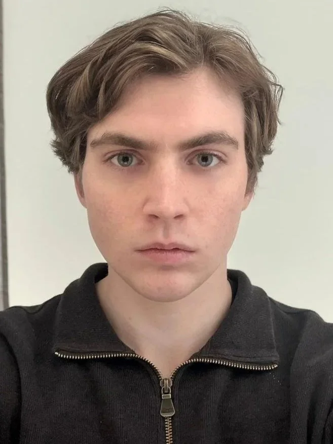 A young man with light skin and medium-length brown hair taking a selfie against a plain light-colored background. He is wearing a black zip-up jacket.