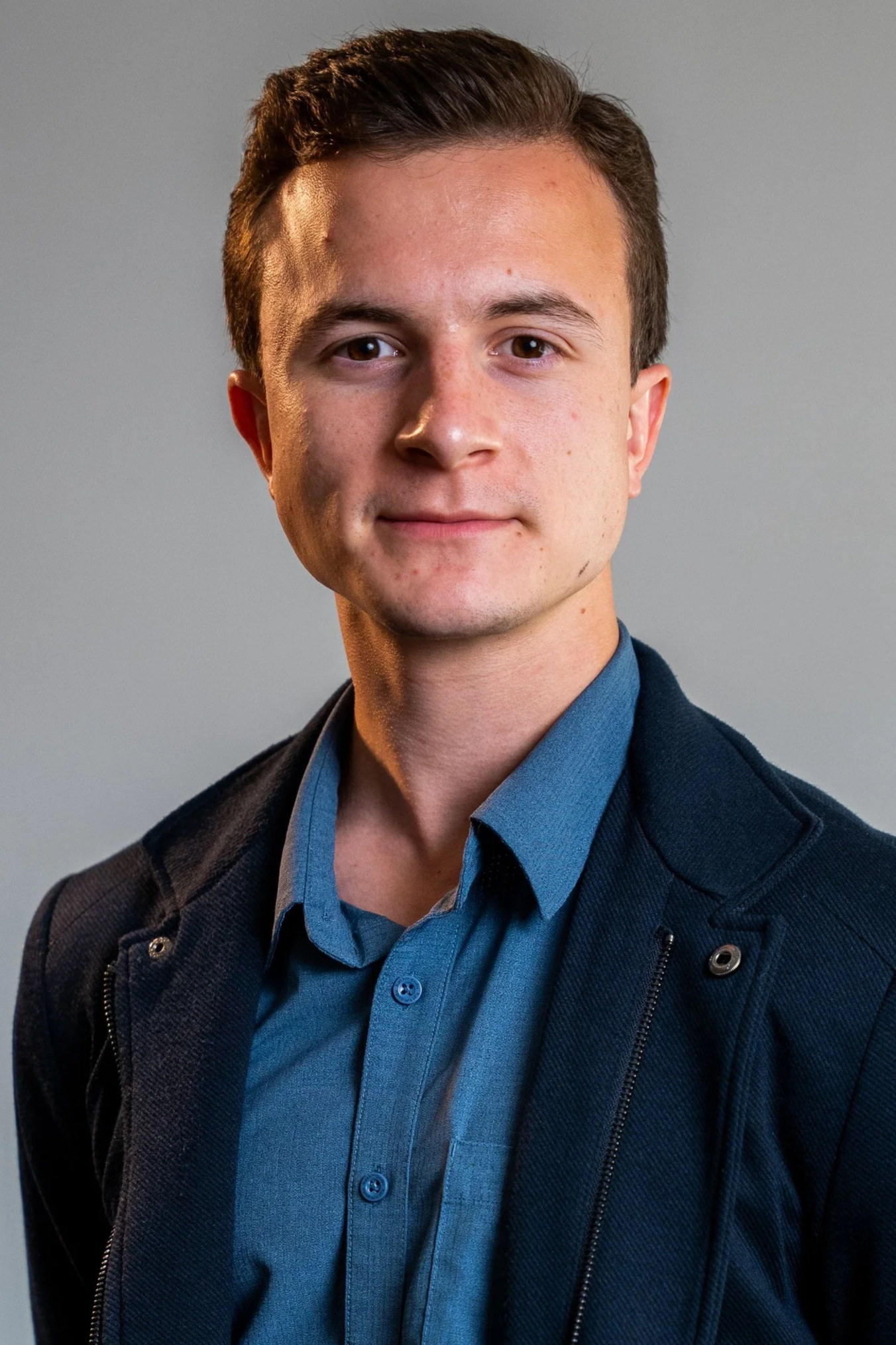 Portrait of a young man with short brown hair, wearing a blue dress shirt and dark blazer, standing in front of a plain gray background.