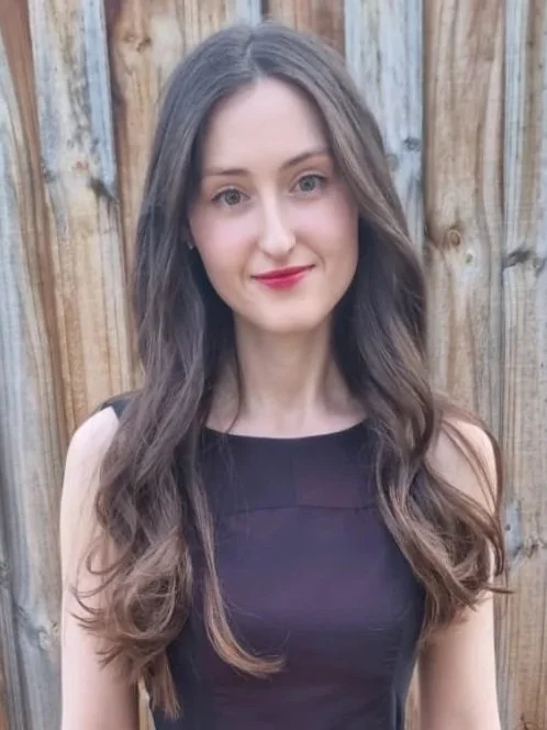 Young woman with long wavy brown hair and fair skin, wearing a sleeveless black top, posing outdoors in front of a wooden fence.