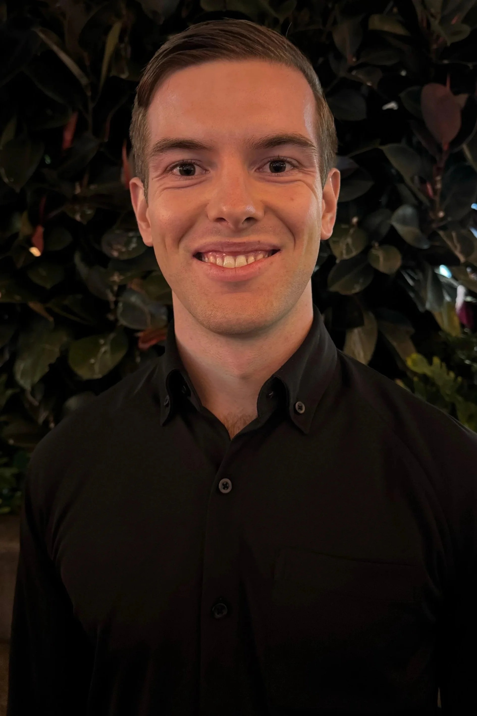A young man with short brown hair and light skin wearing a black button-up shirt, smiling, in front of a backdrop of leafy plants.