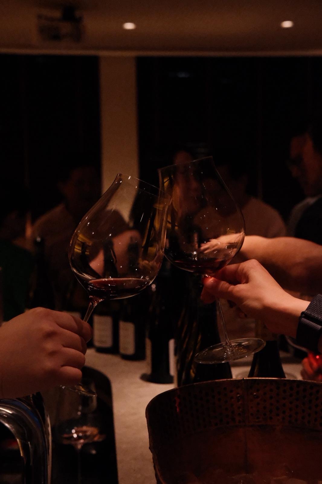 Two people clinking glasses of red wine at a dimly lit gathering or celebration.