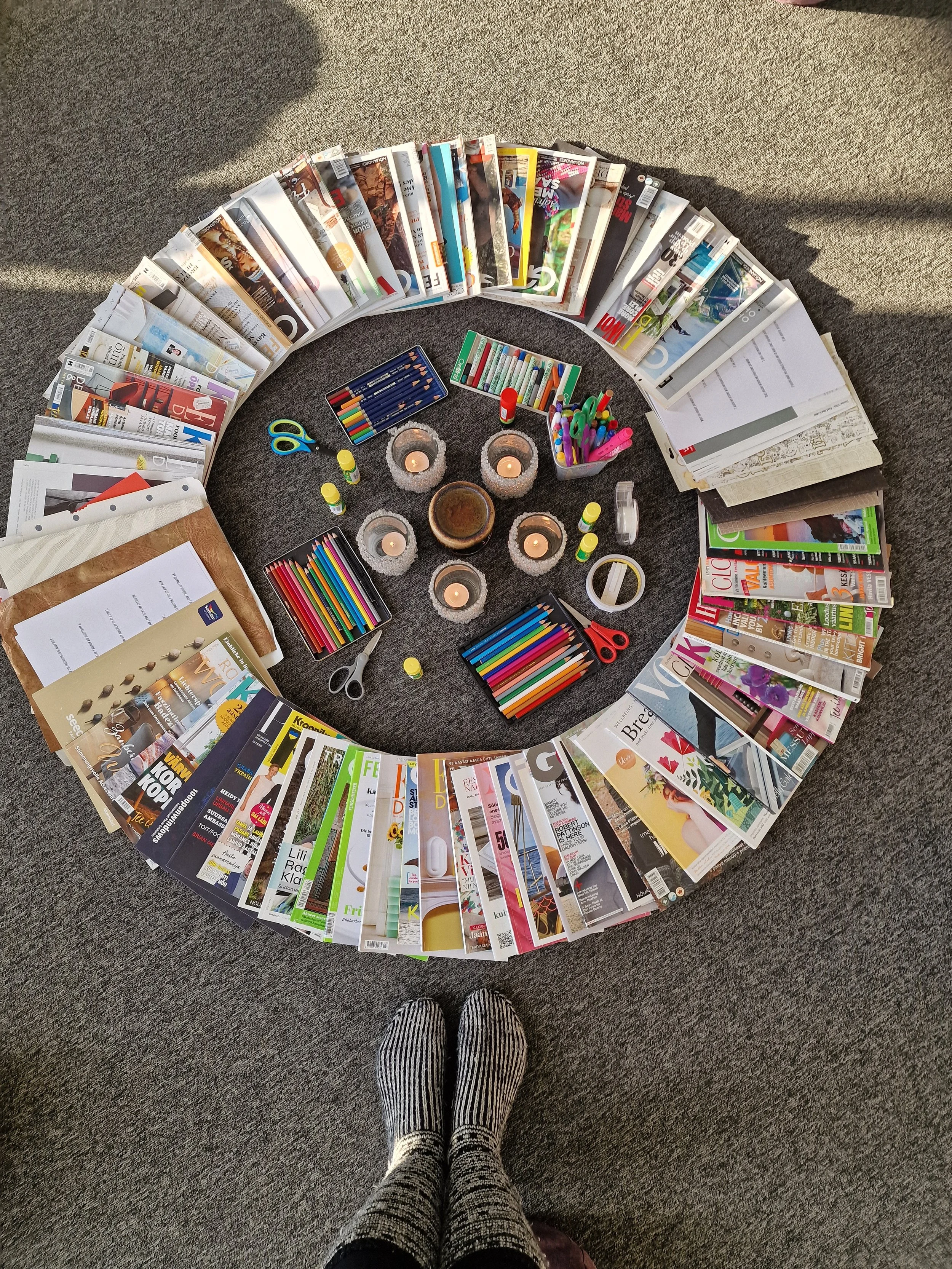 A circular arrangement of magazines on a gray carpeted floor, with craft supplies like scissors, scissors, glue sticks, markers, and candles in the center, and a person's feet in striped socks visible at the bottom of the image.