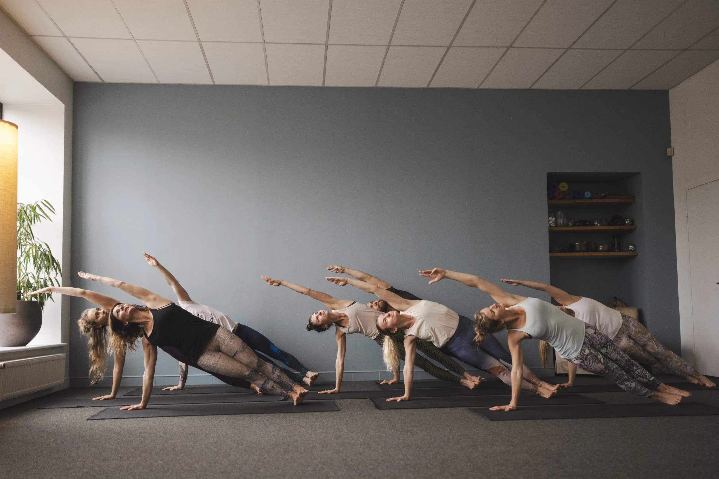 Yoga practice in a studio, performing a side plank pose with one hand on the ground and the other arm extended upwards, on black yoga mats.