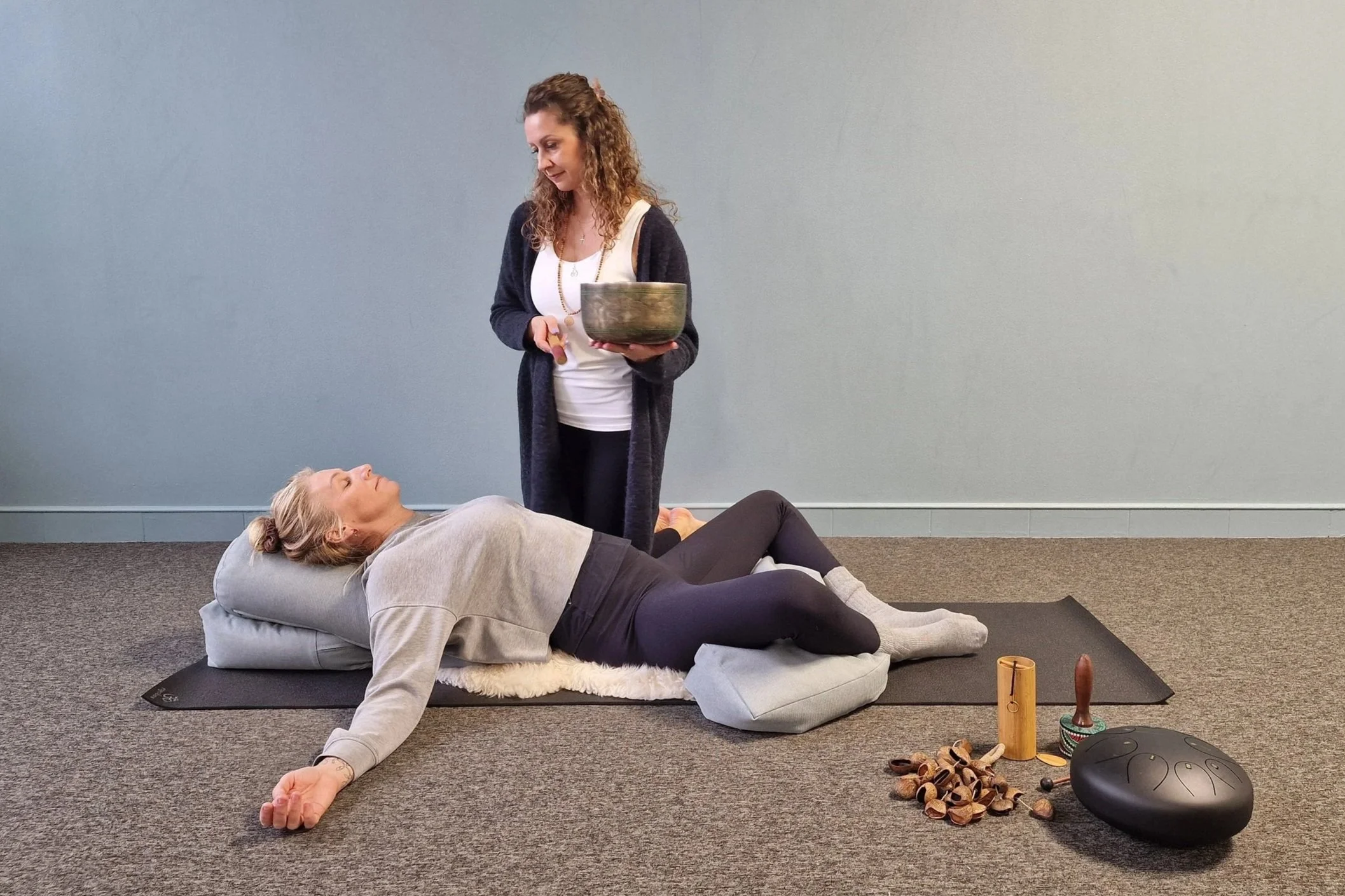 A woman lying on her side on a yoga mat, with a knee pillow, resting her head on a pillow, as another woman standing nearby holds a tibetan singing bowl. There are musical shakers, a mallet, and a black singing bowl on the floor next to them.