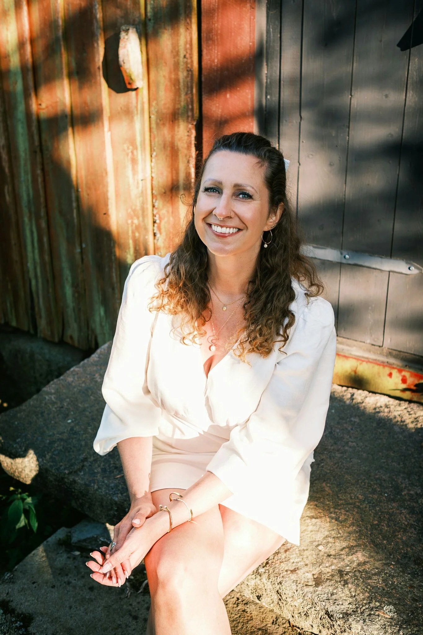 A woman with curly brown hair, wearing a white dress, sitting on a rock outdoors, smiling, with a rustic wooden and metal background.