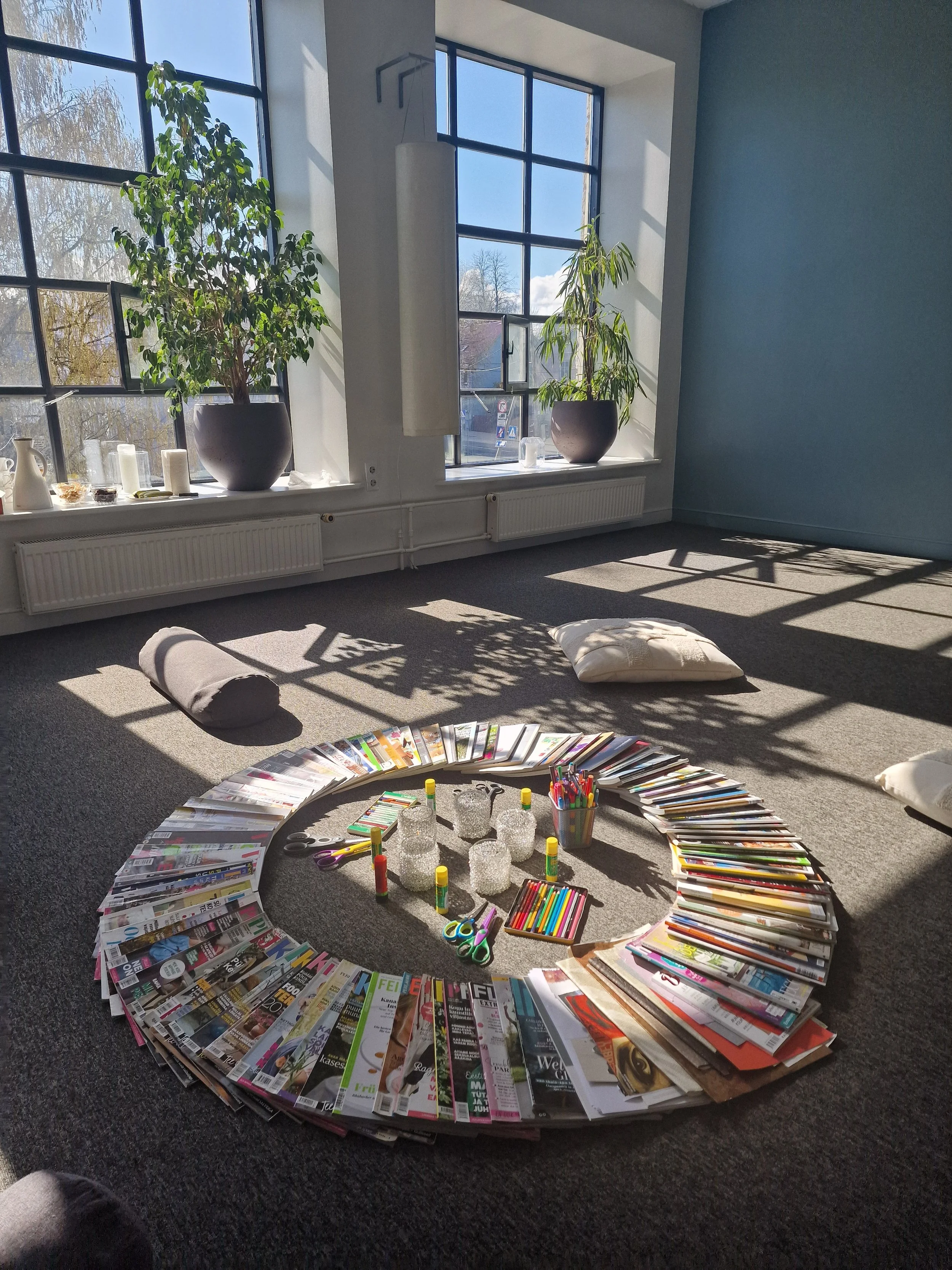Sunlit room with large windows, two potted plants on the windowsill, and a circular arrangement of magazines, colored markers, glue sticks, and craft supplies on the carpeted floor.