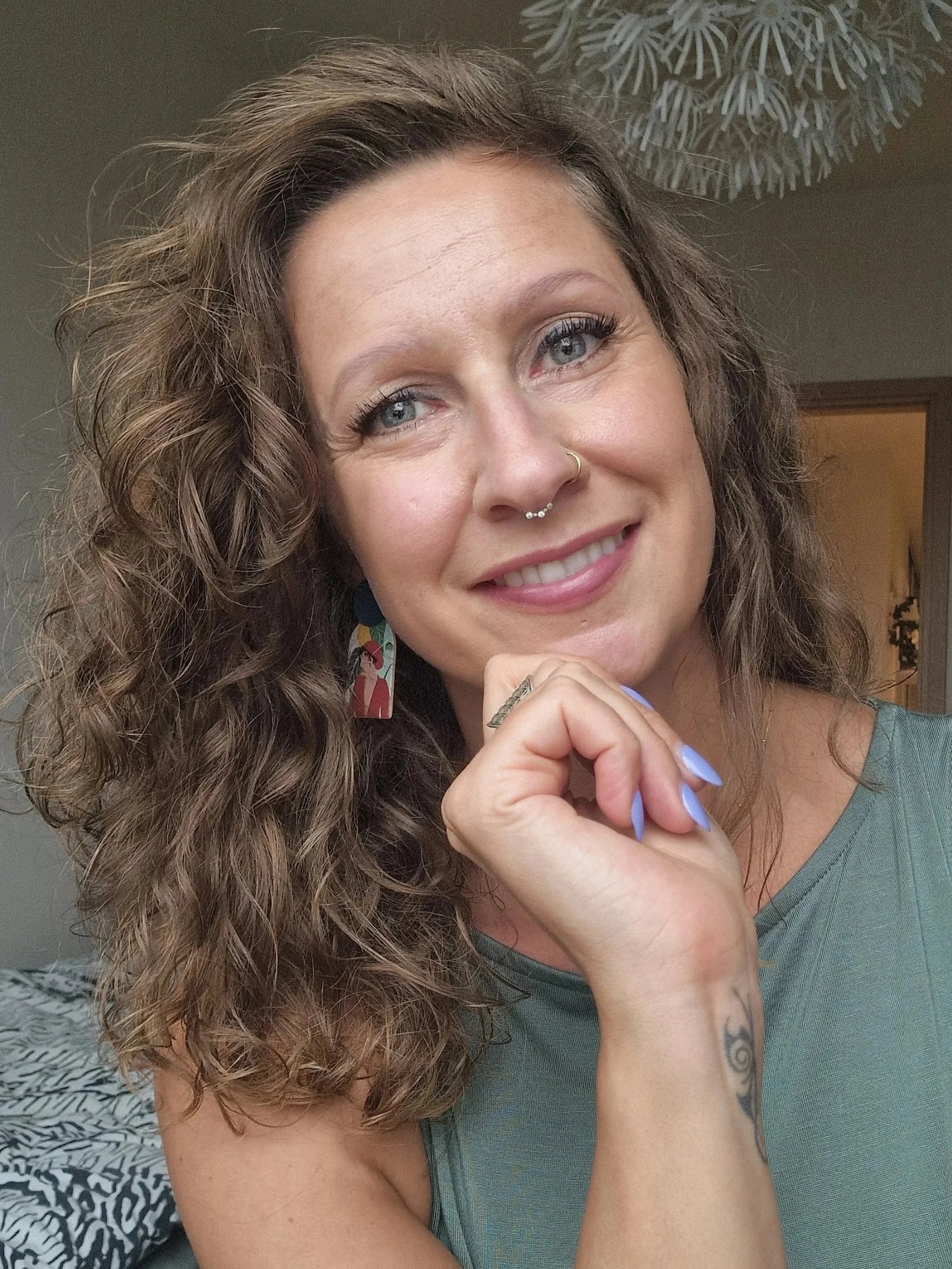 A woman with curly brown hair, blue eyes, and a septum piercing, smiling and resting her chin on her hand, wearing a green top.