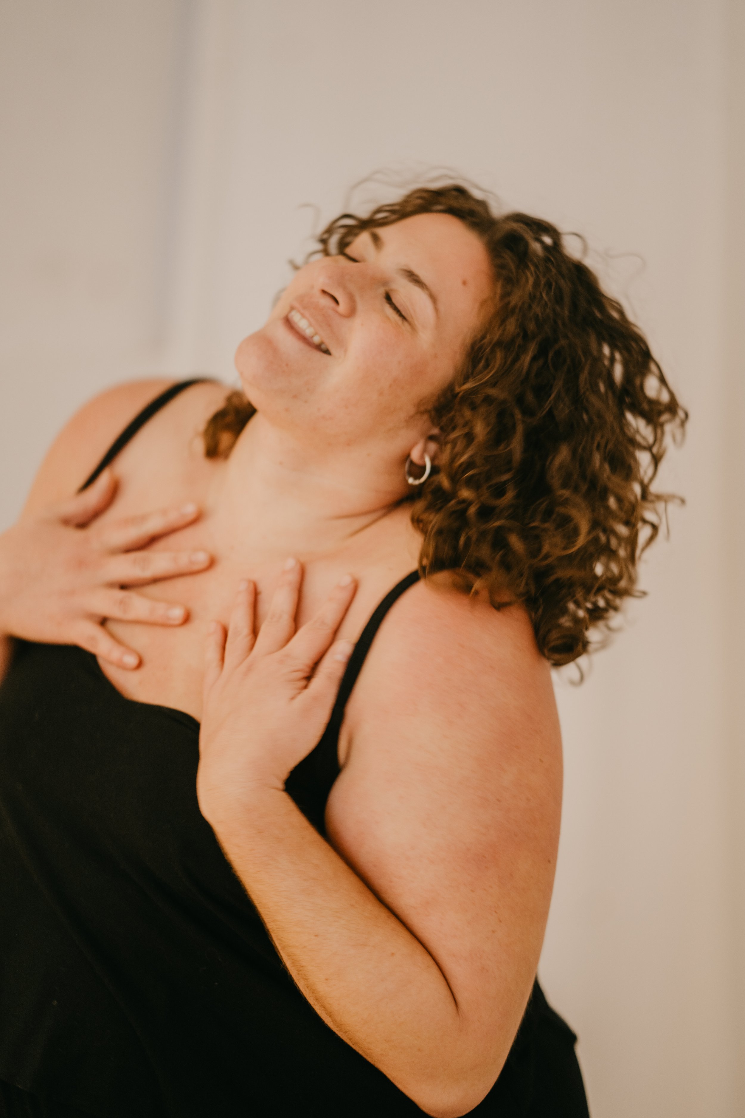 Mujer con cabello rizado y sonrisa, con las manos en el pecho y los ojos cerrados.