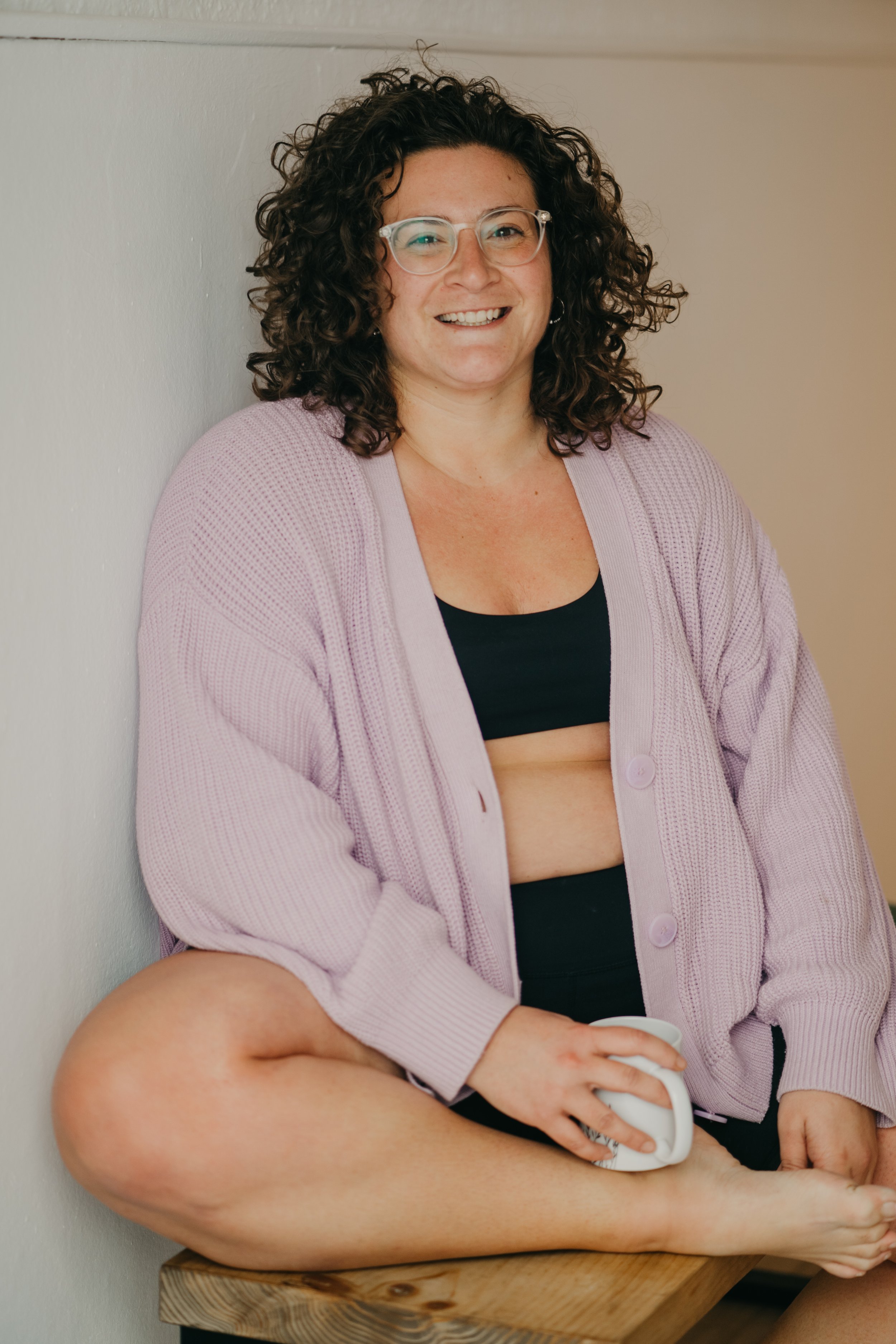 Mujer sonriendo, con gafas y cabello rizado, usando un suéter rosado, sosteniendo una taza en una mano, sentada en un banco de madera.