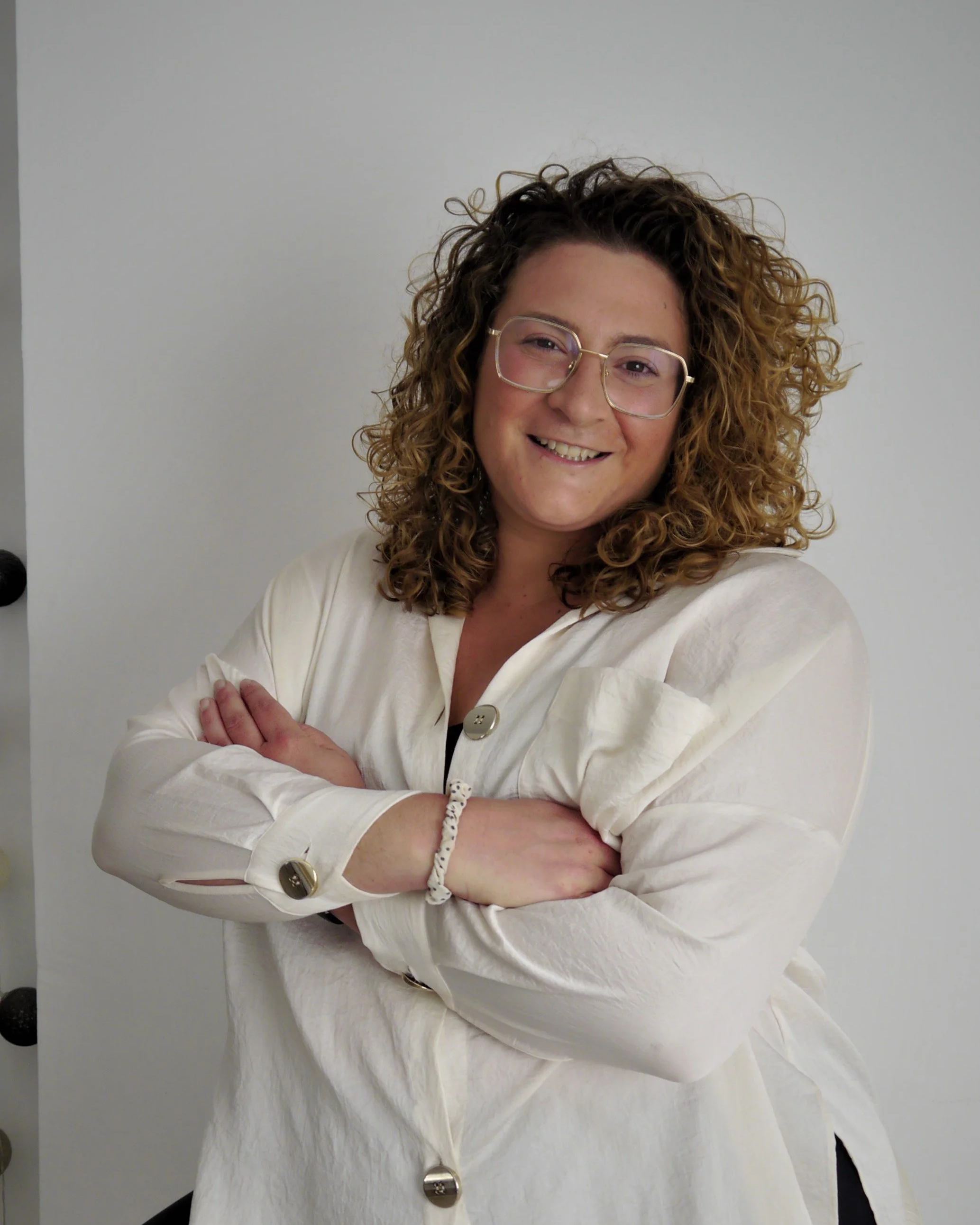 Mujer con cabello rizado y lentes, sonriendo con brazos cruzados, vestida con blusa blanca y fondo gris.