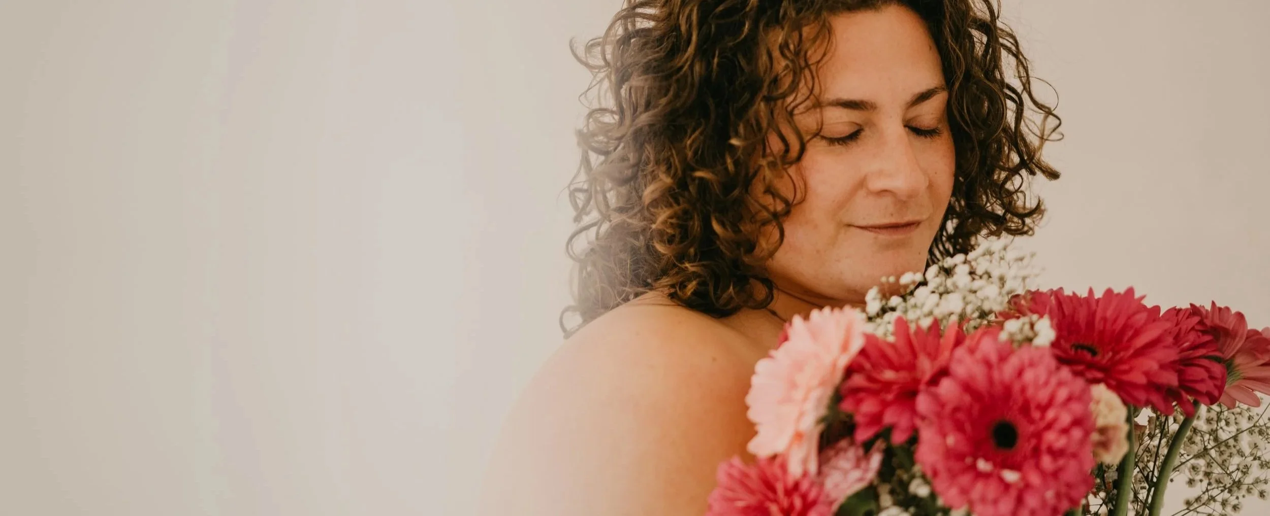 Mujer con cabello rizado y cerrar ojos sosteniendo un ramo de flores rojas, rosadas y blancas, con fondo neutro.