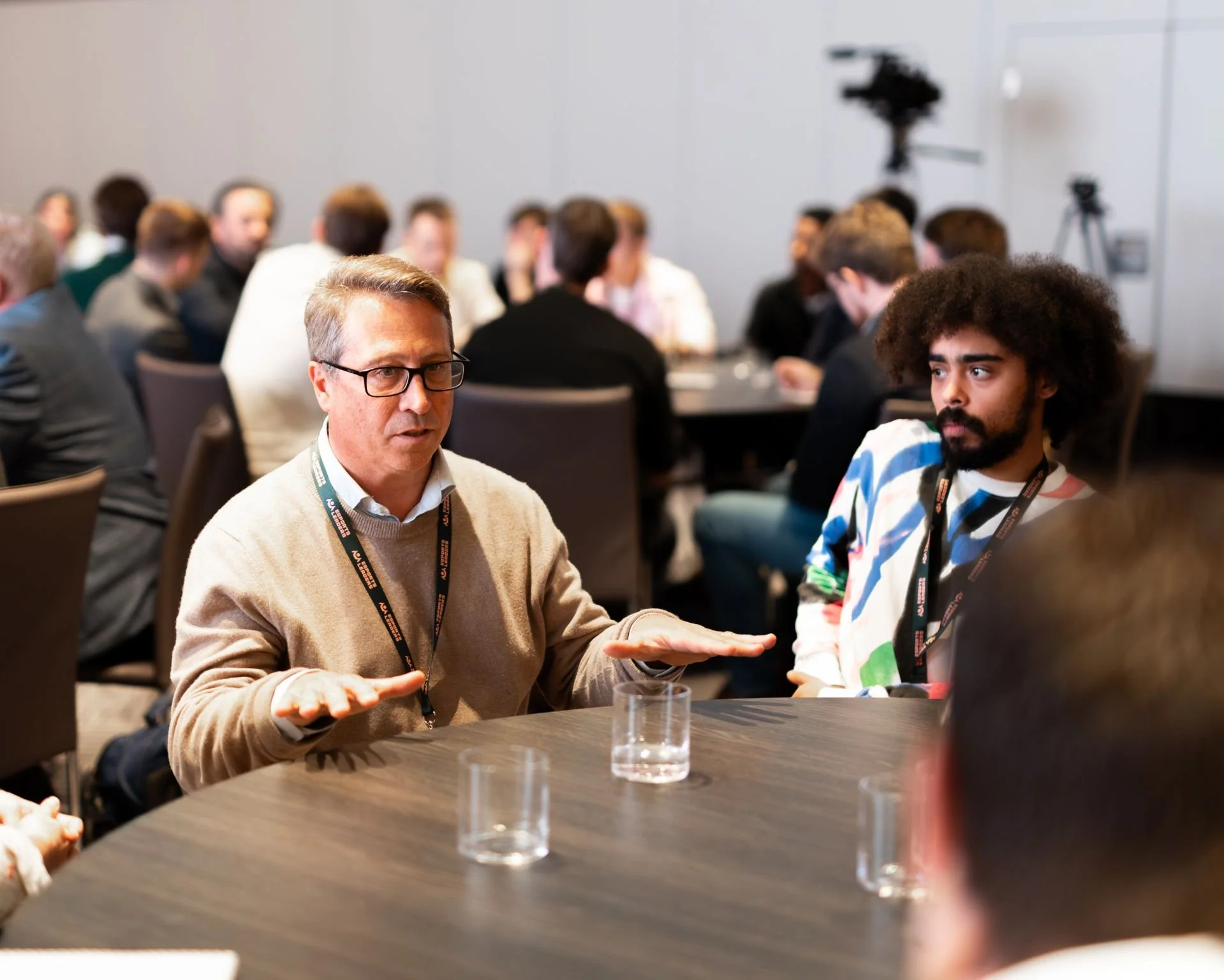 A group of people in a conference room, with two individuals in the foreground engaged in conversation, one gesturing with his hands.