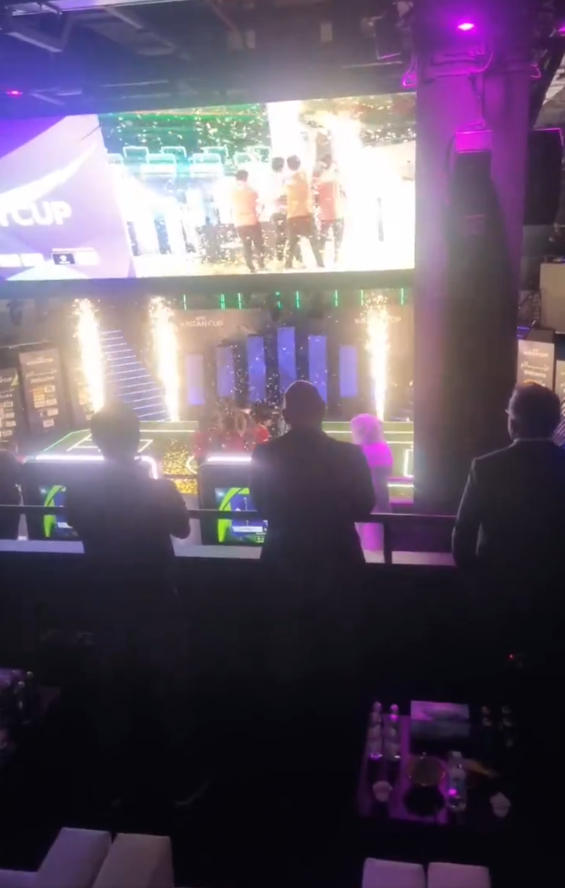 People watching a large screen displaying a scene from a sports event in a dark, nightclub-like venue with neon lighting and fireworks.