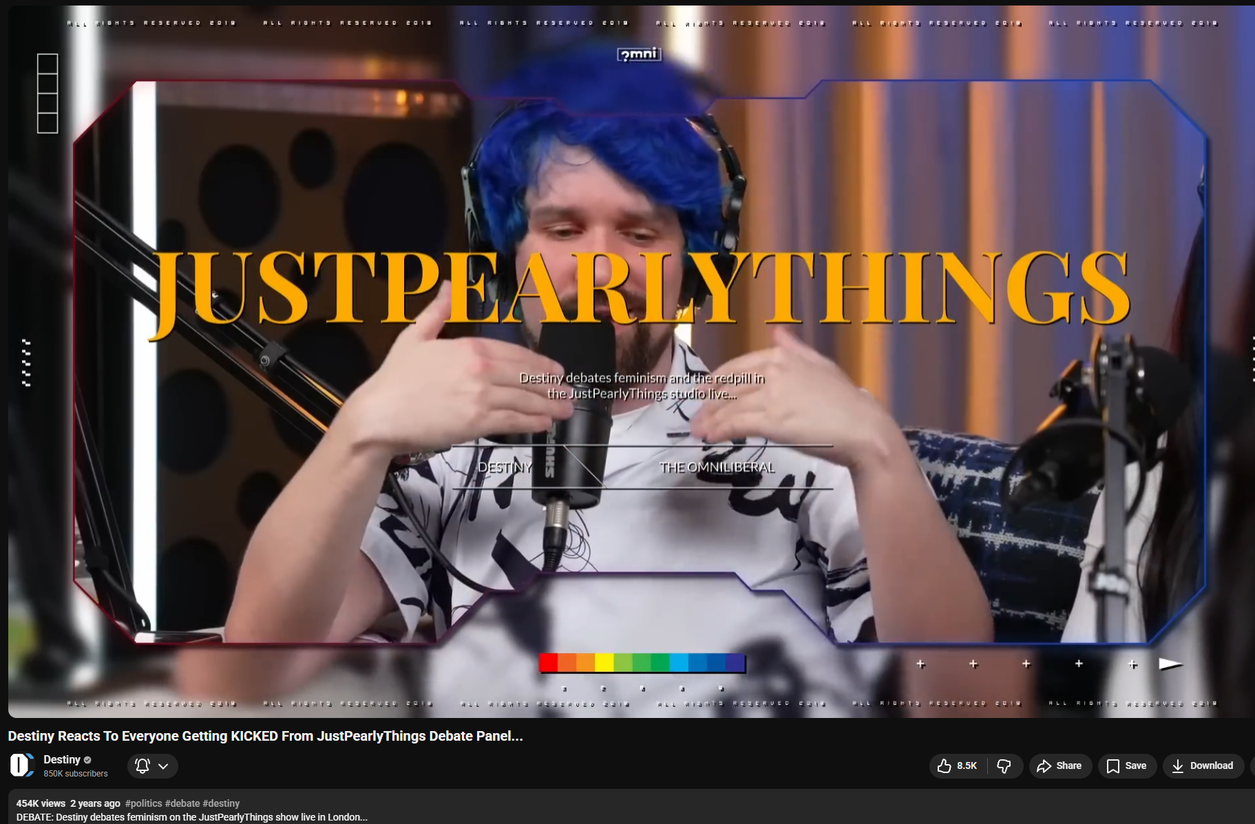 Person with blue hair and headphones speaking into a microphone during a recorded podcast or debate, with overlay text 'JustPearlyThings' and context about the debate topic.
