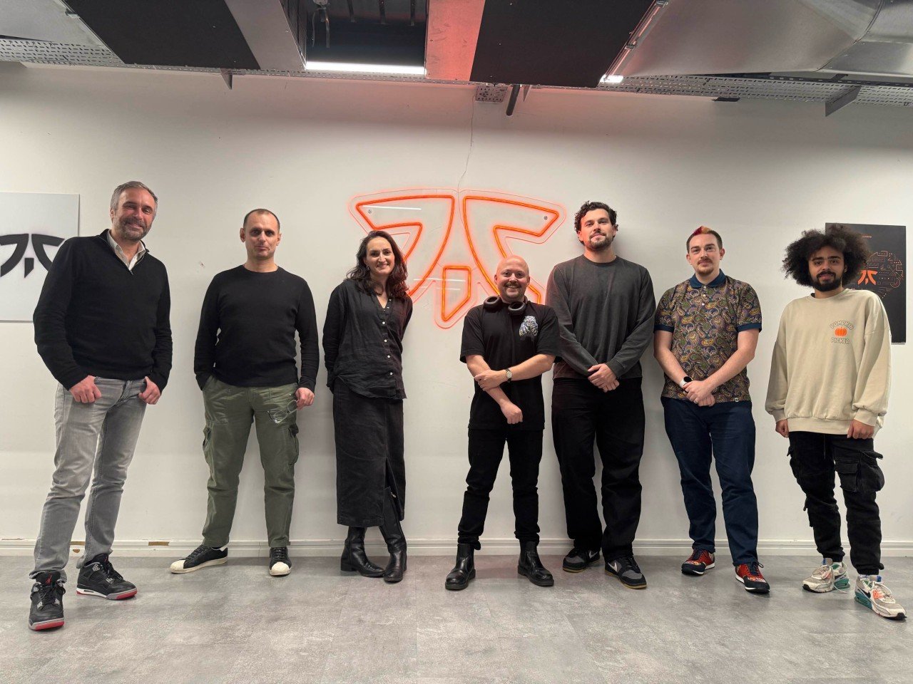 Seven people standing in front of a white wall with an illuminated orange logo, posing for a group photo indoors.