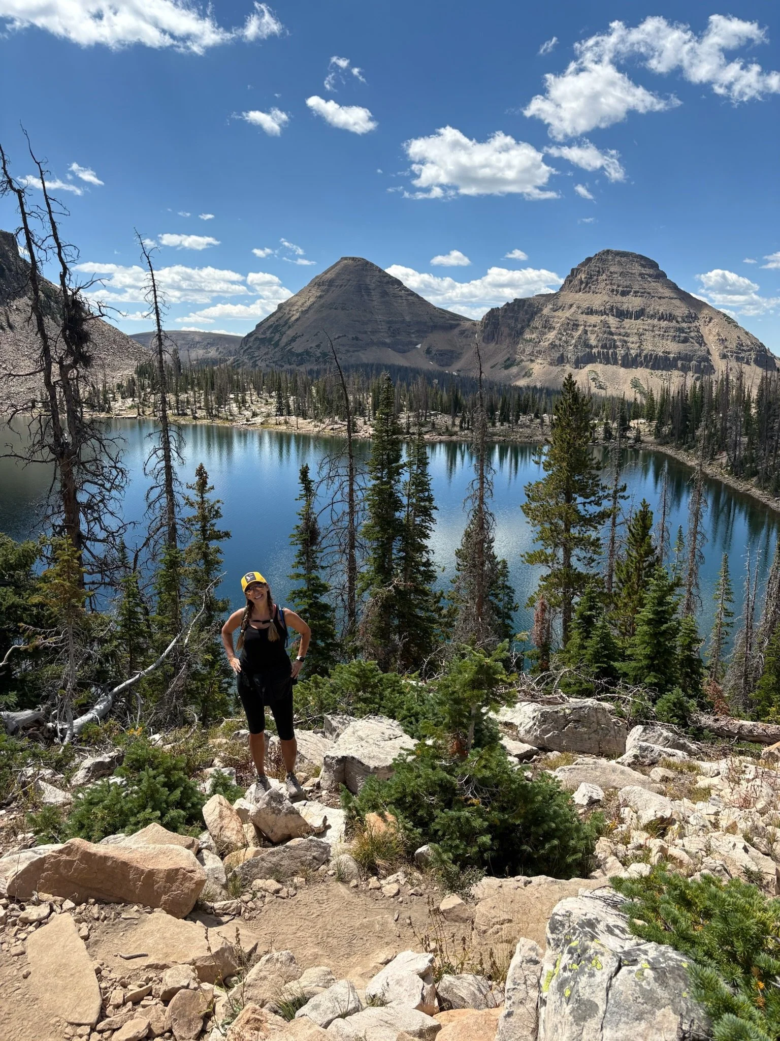 A woman standing on rocky terrain near a large mountain lake with evergreen trees, mountain peaks, and partly cloudy sky in the background.