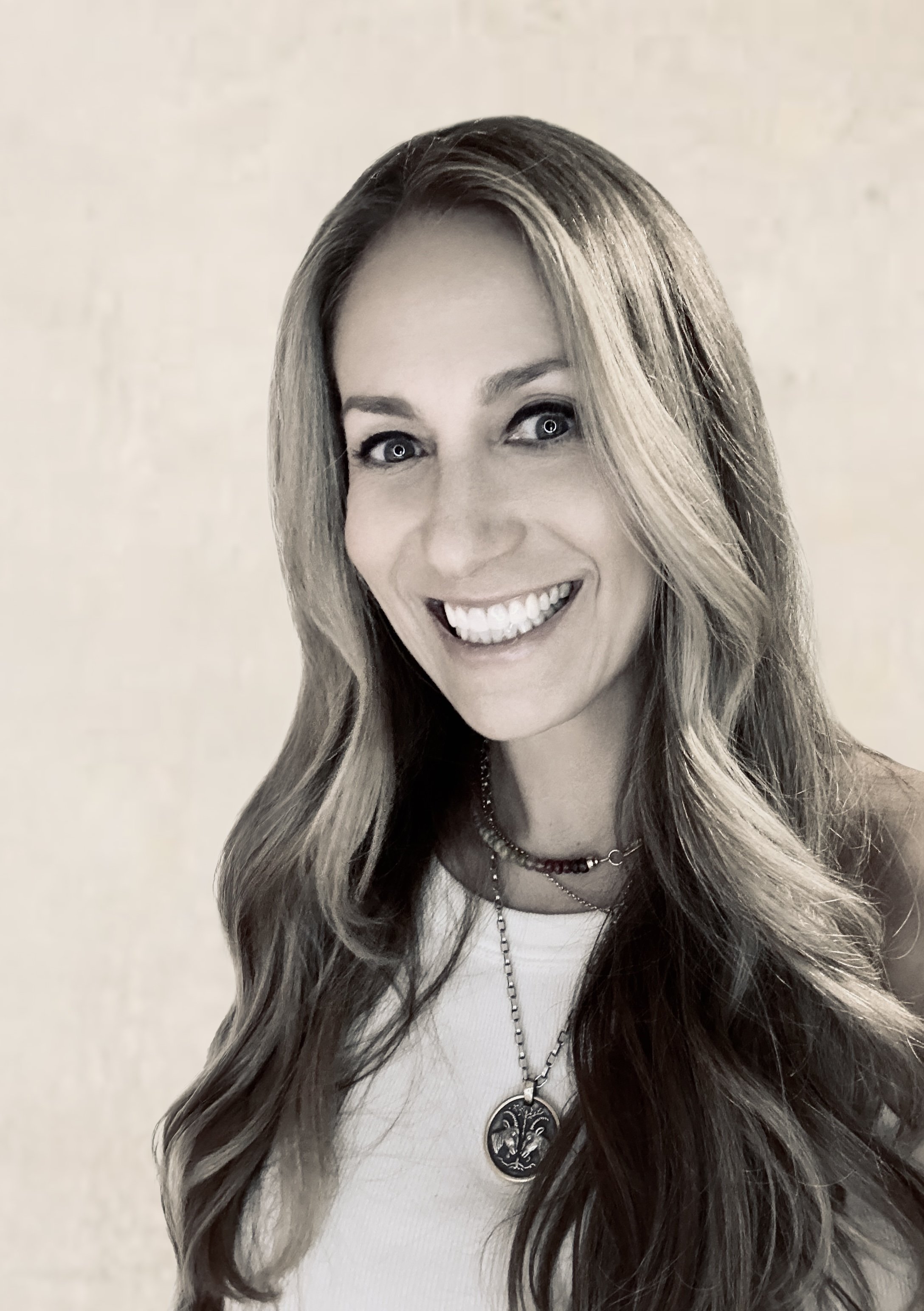 A smiling woman with long wavy hair wearing layered necklaces and a sleeveless top.