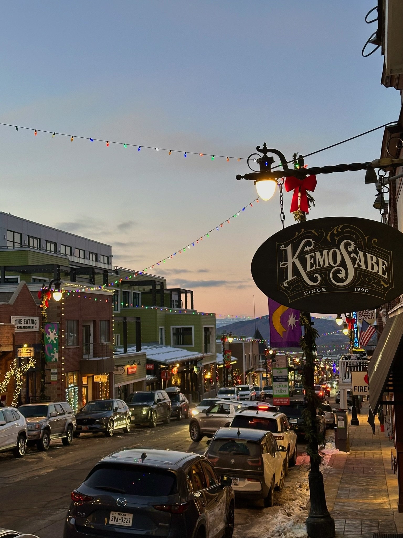 Street scene at dusk with parked cars and buildings decorated with Christmas lights and ornaments, including a sign for a restaurant or shop called 'Kemo Sabe' and colorful string lights overhead.