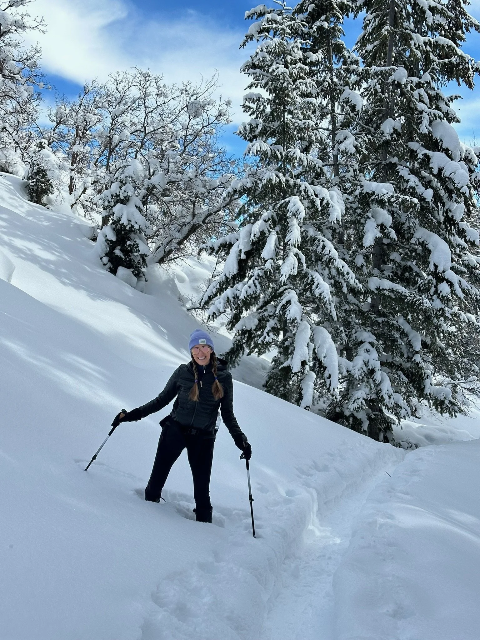 A woman with glasses and a purple hat hiking in snowy landscape with snow-covered trees and blue sky.