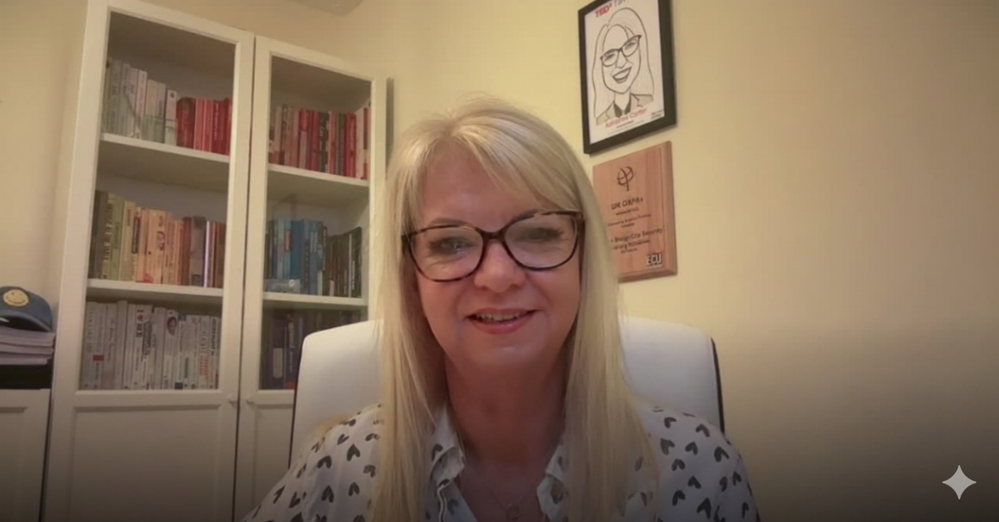 A blonde woman wearing glasses and a white shirt with black hearts sitting in a room with bookshelves and framed pictures on the wall.