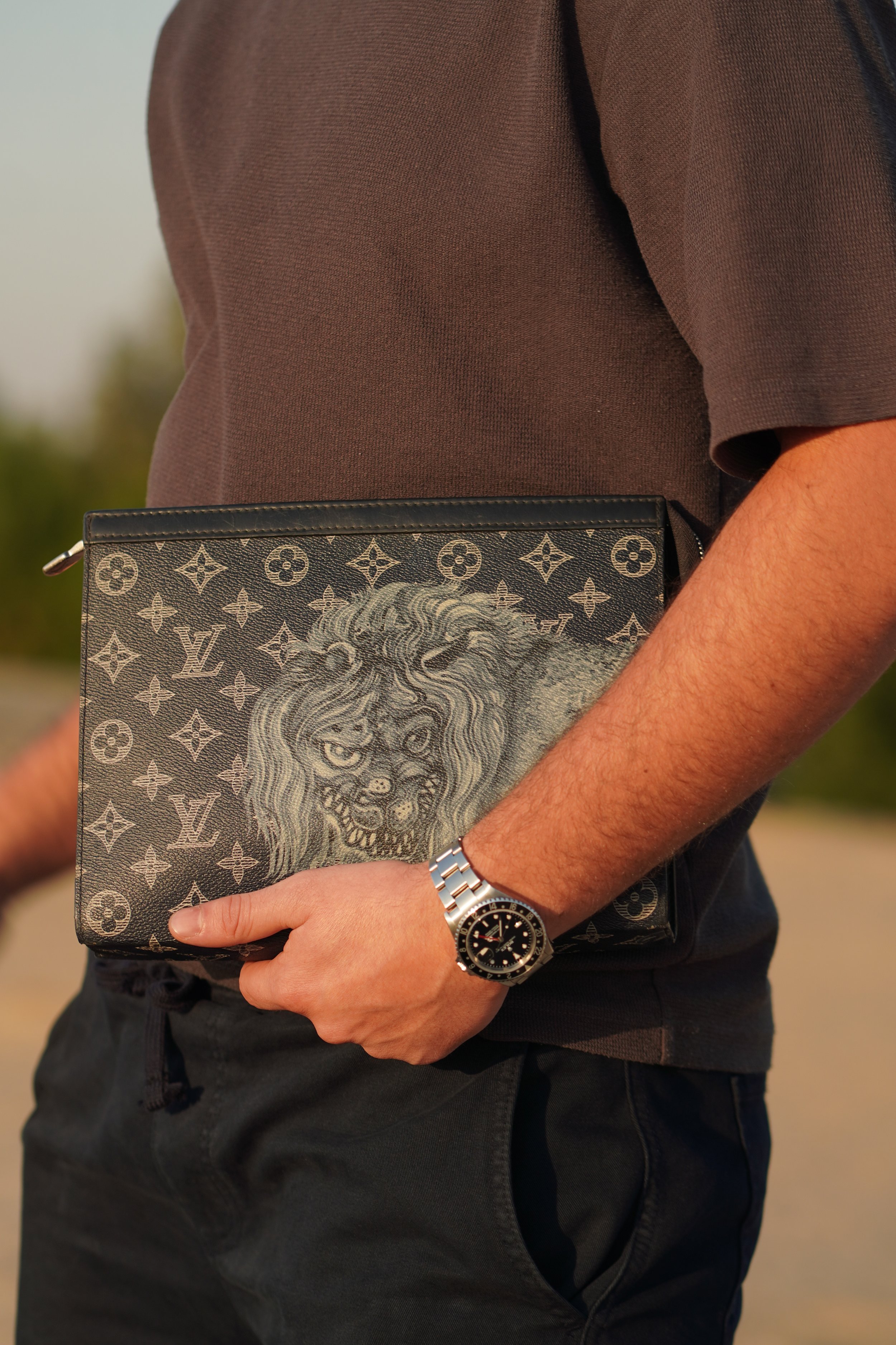 A person wearing a gray shirt and black pants holding a Louis Vuitton clutch with a lion's face graphic and a background pattern of Louis Vuitton monograms, while wearing a wristwatch.