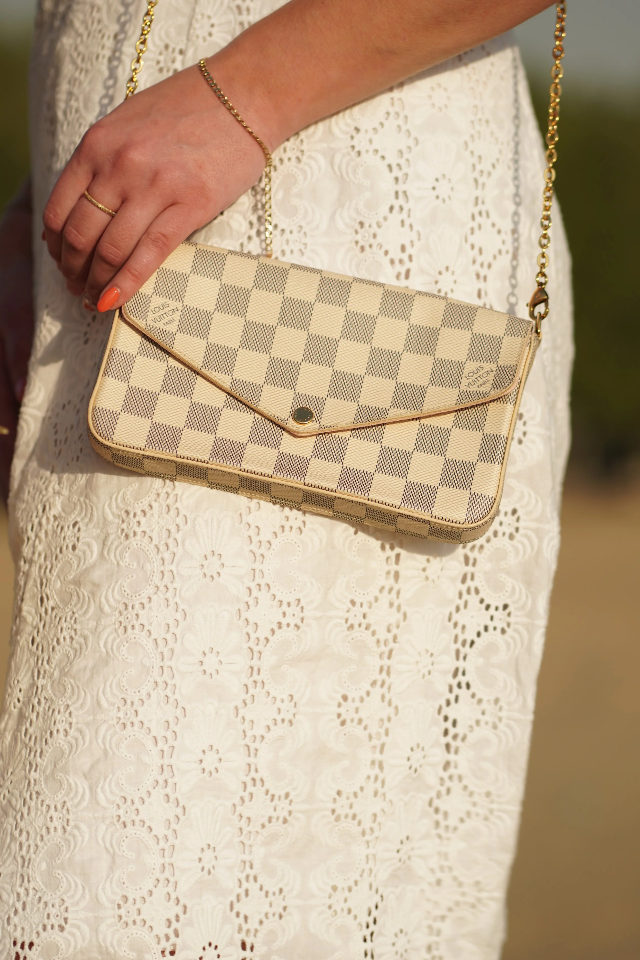 A person wearing a cream-colored, intricately embroidered dress holding a beige Louis Vuitton checkered clutch with a gold chain strap.