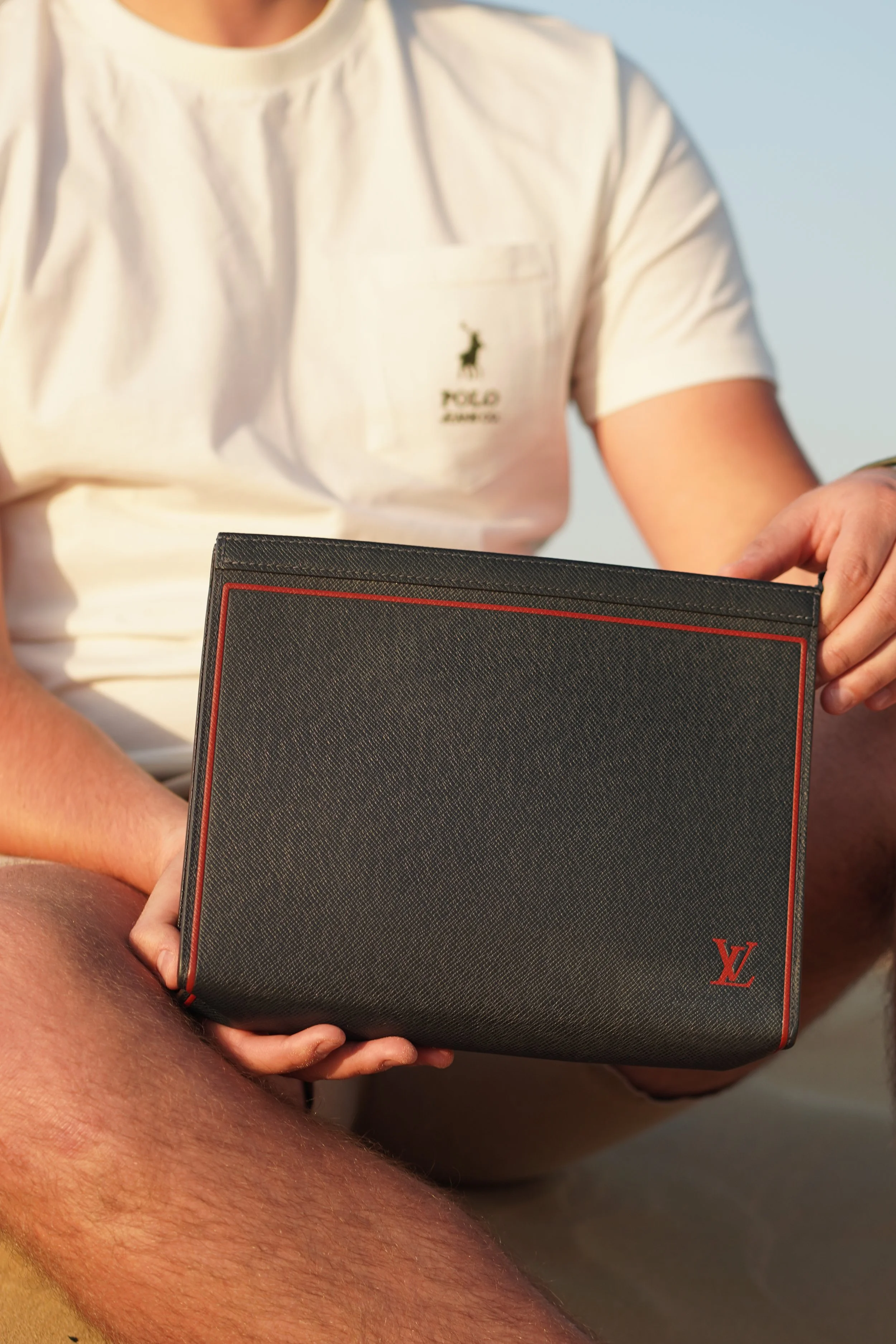 A person in a white Polo shirt holding a black Louis Vuitton clutch with red accents outdoors.