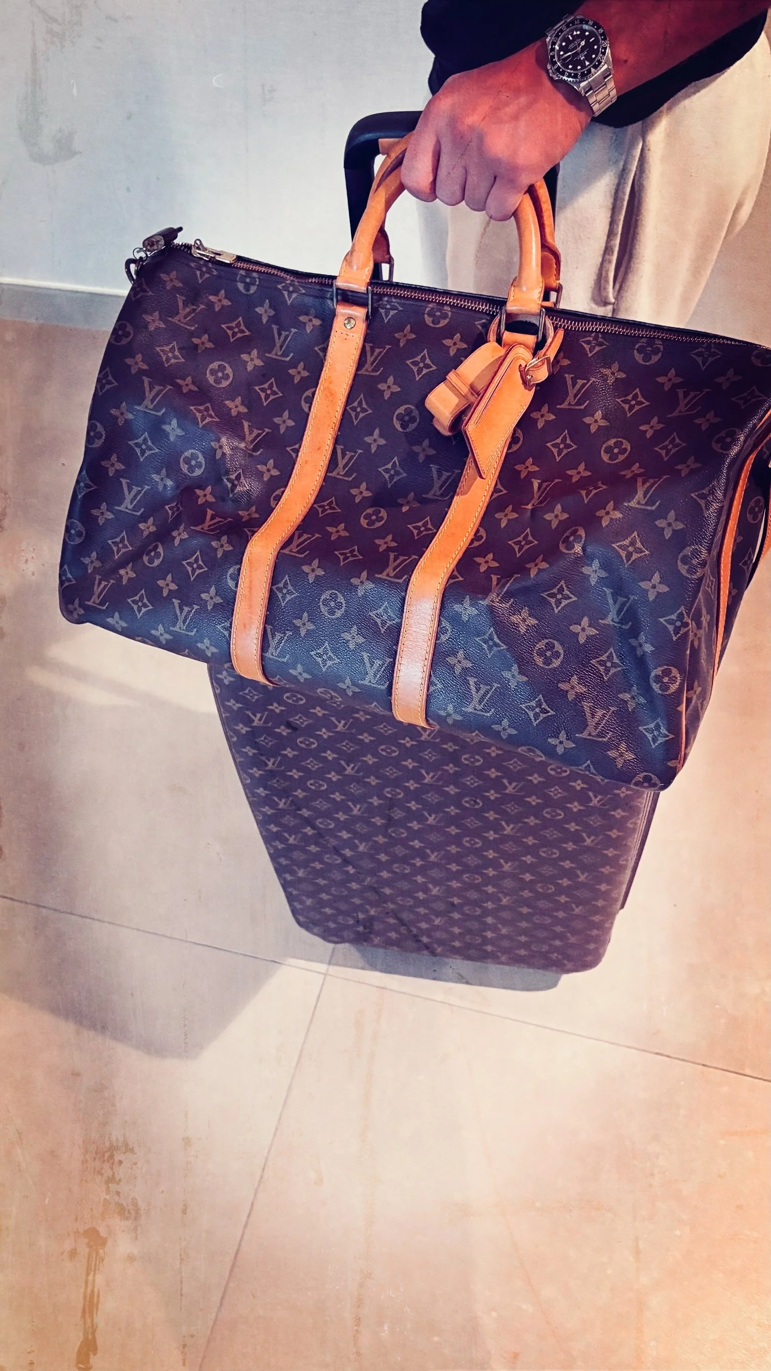 Person holding a Louis Vuitton monogram canvas duffel bag with tan leather handles and trim, standing on a tiled floor.