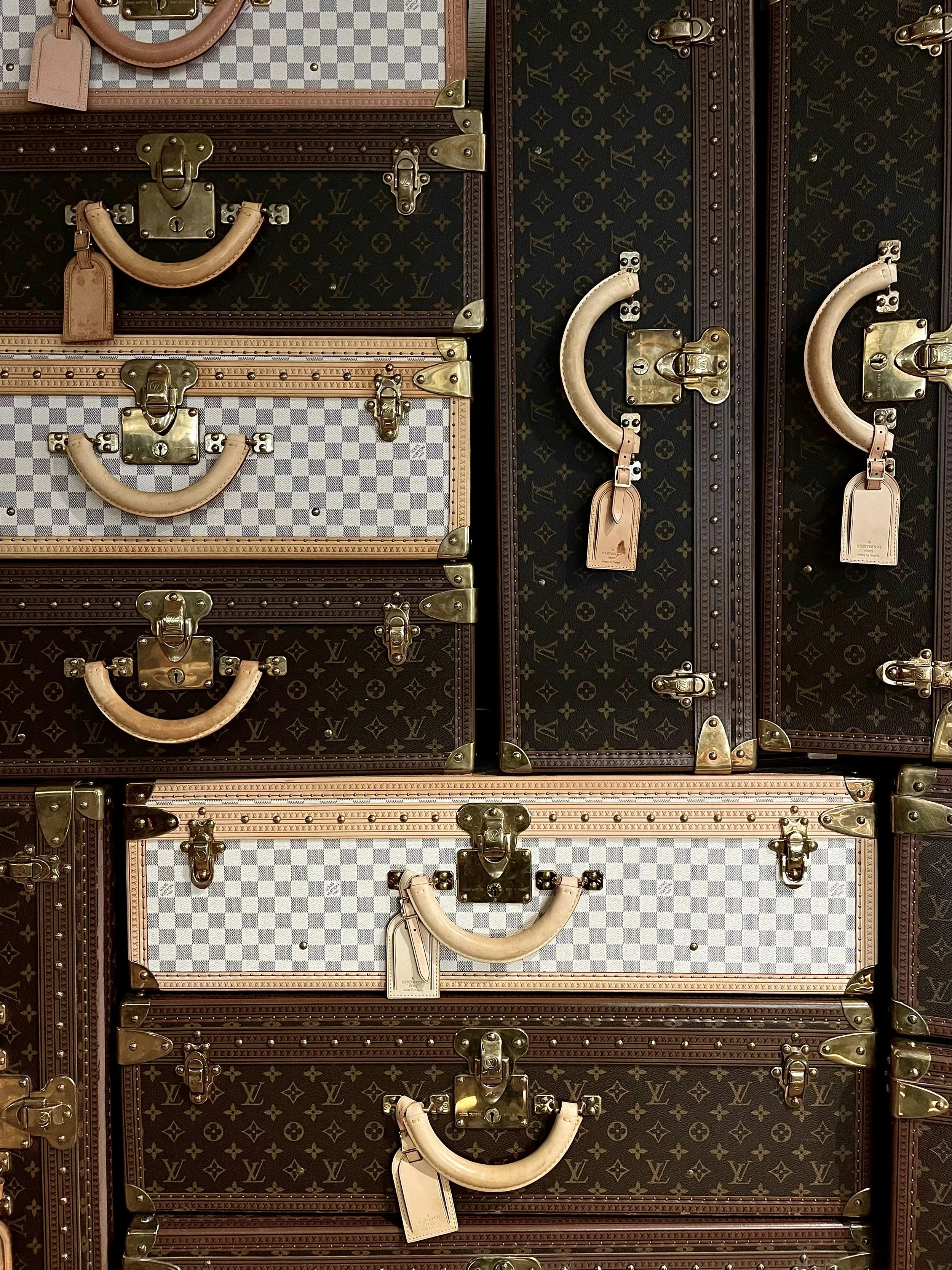 Multiple vintage Louis Vuitton suitcases stacked on top of each other, featuring various monogram and checkered patterns, with leather handles and brass locks.