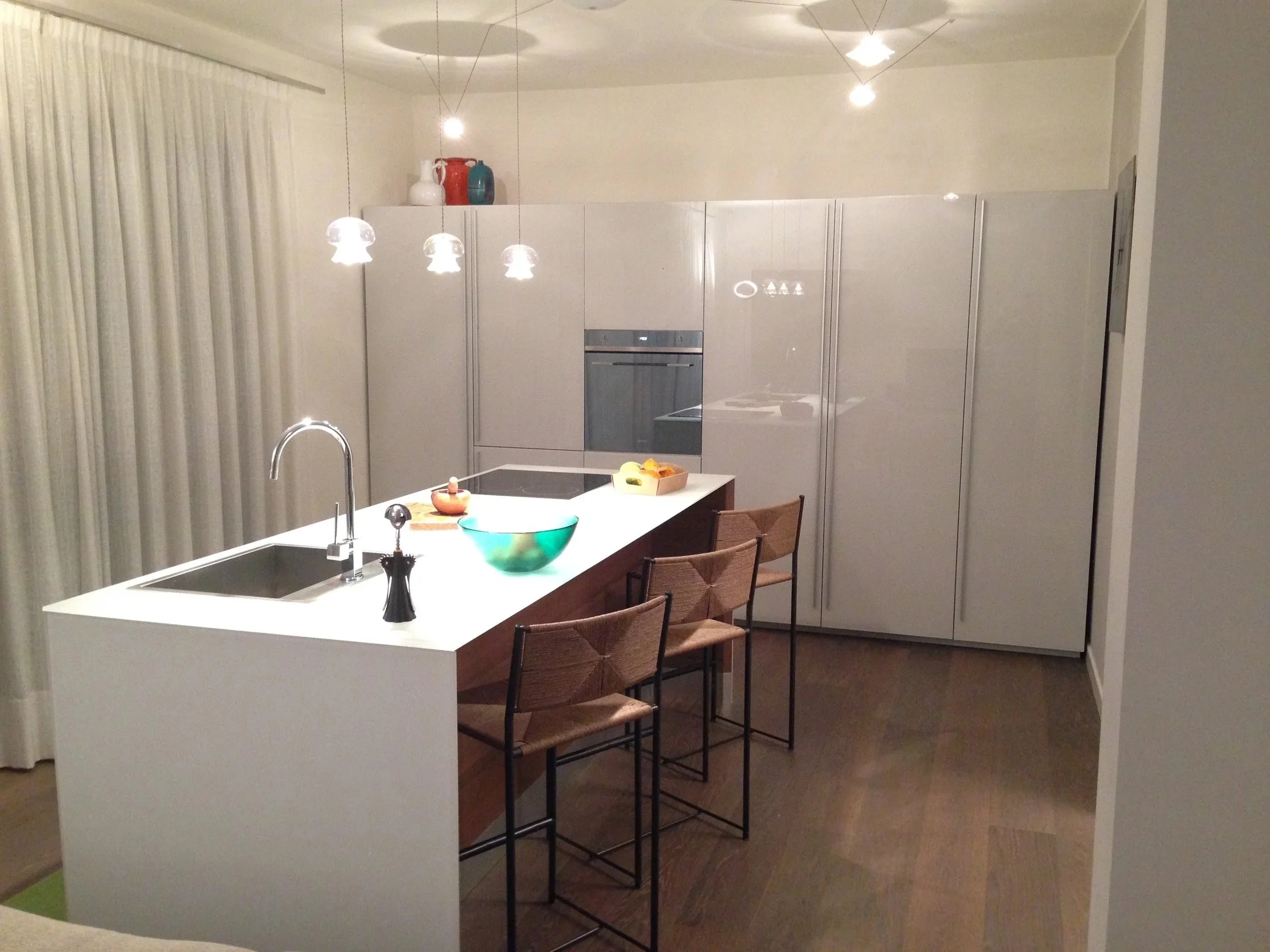 Cucina moderna con isola centrale bianca e tre sedie in ferro e legno, armadi bianchi, pavimento in legno, illuminazione a led, decorazioni in alto e oggetti vari sulla superficie dell'isola.